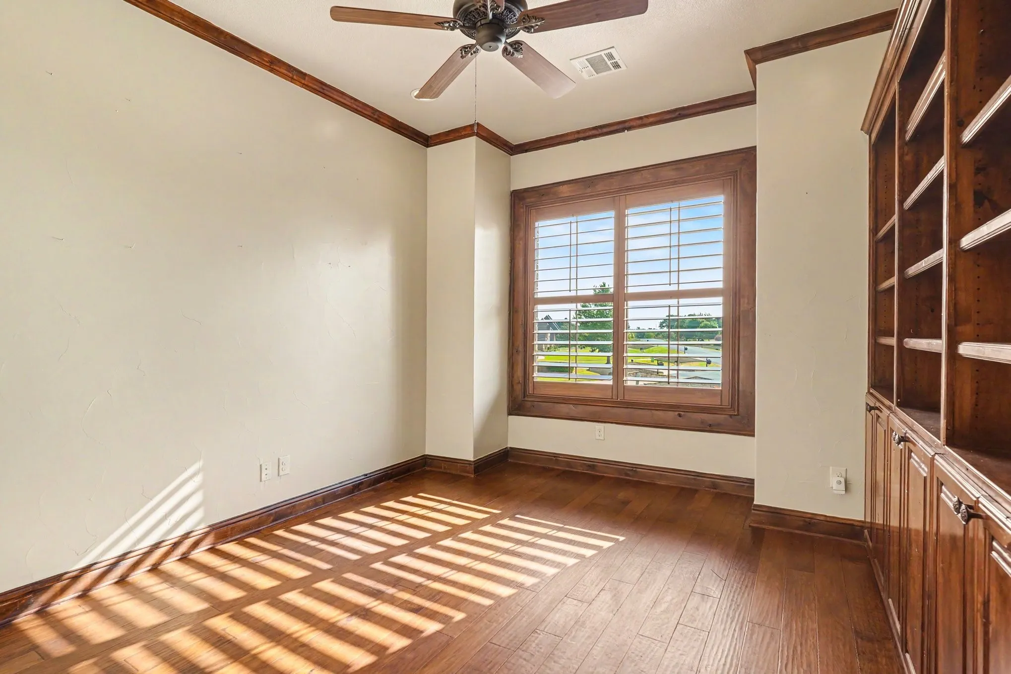 Wood-finish flooring, built-in wooden shelving, window with wood shutters, wood trim, and a ceiling fan