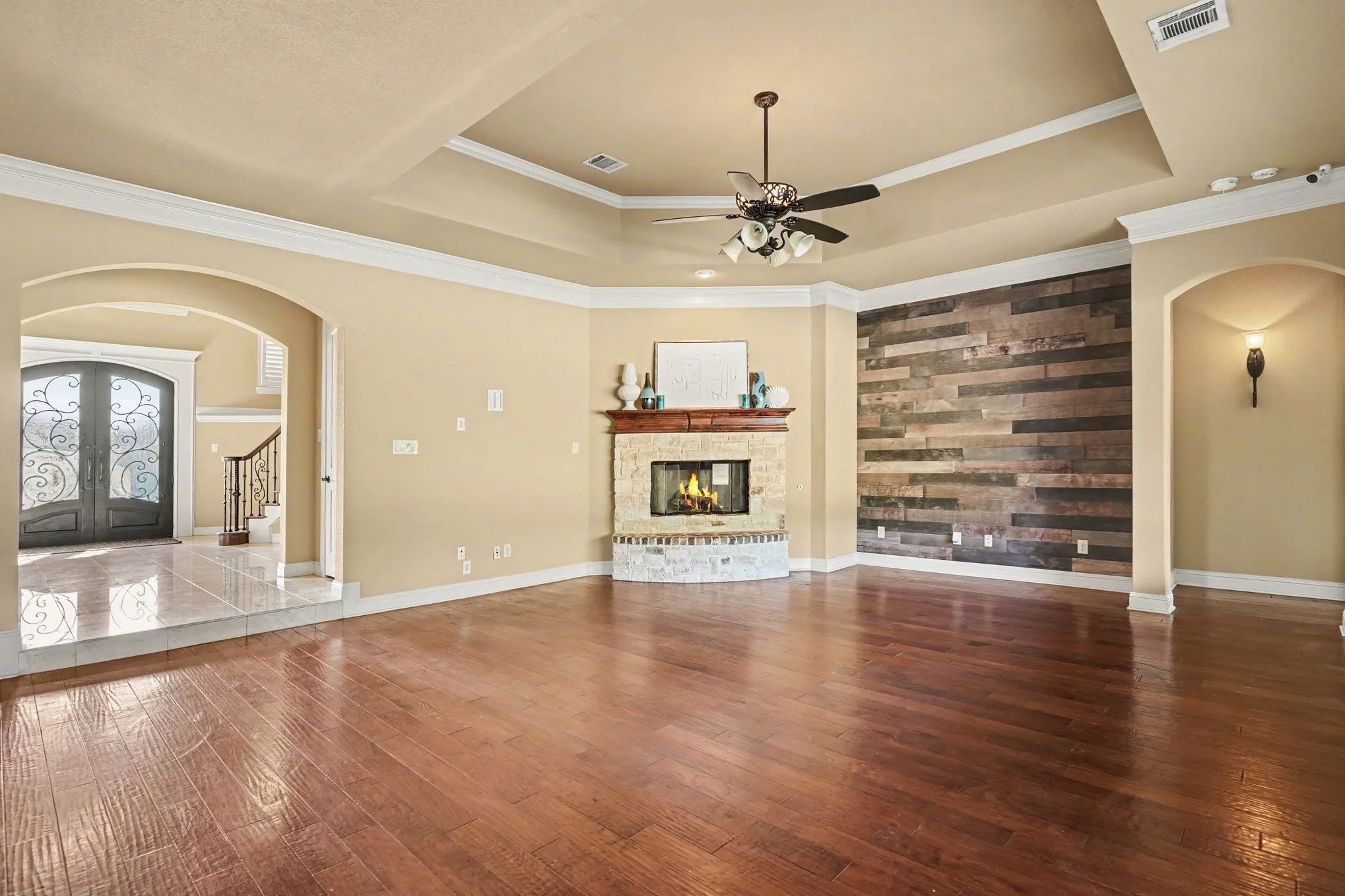 Tray ceiling with crown molding, wood-finish flooring, stone-clad fireplace with mantel, accent wall with wood paneling, and arched doorways