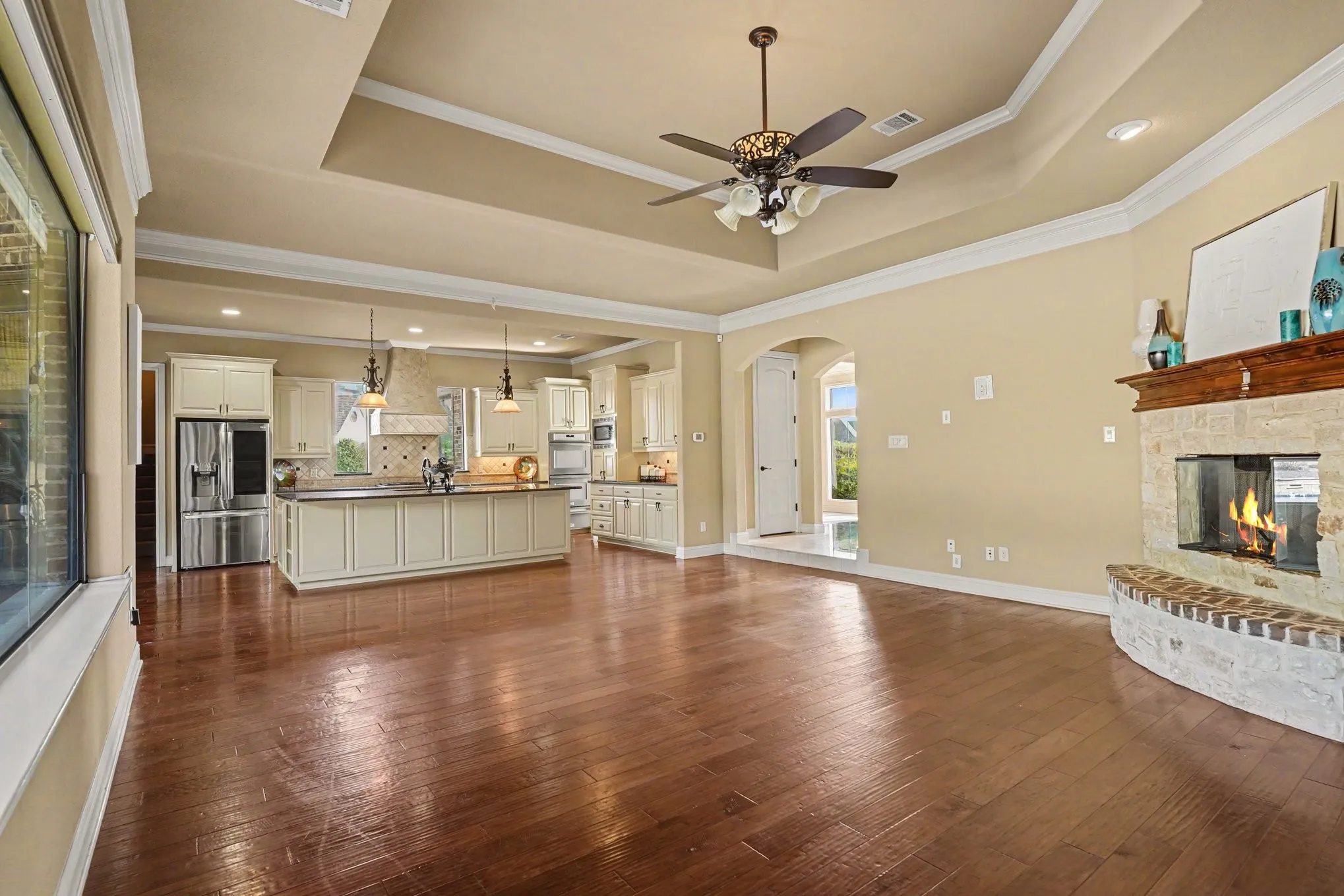 Wood-finish flooring throughout, tray ceiling with a ceiling fan, stone-surround fireplace with a wood mantle, recessed lighting, and a kitchen island with pendant lighting