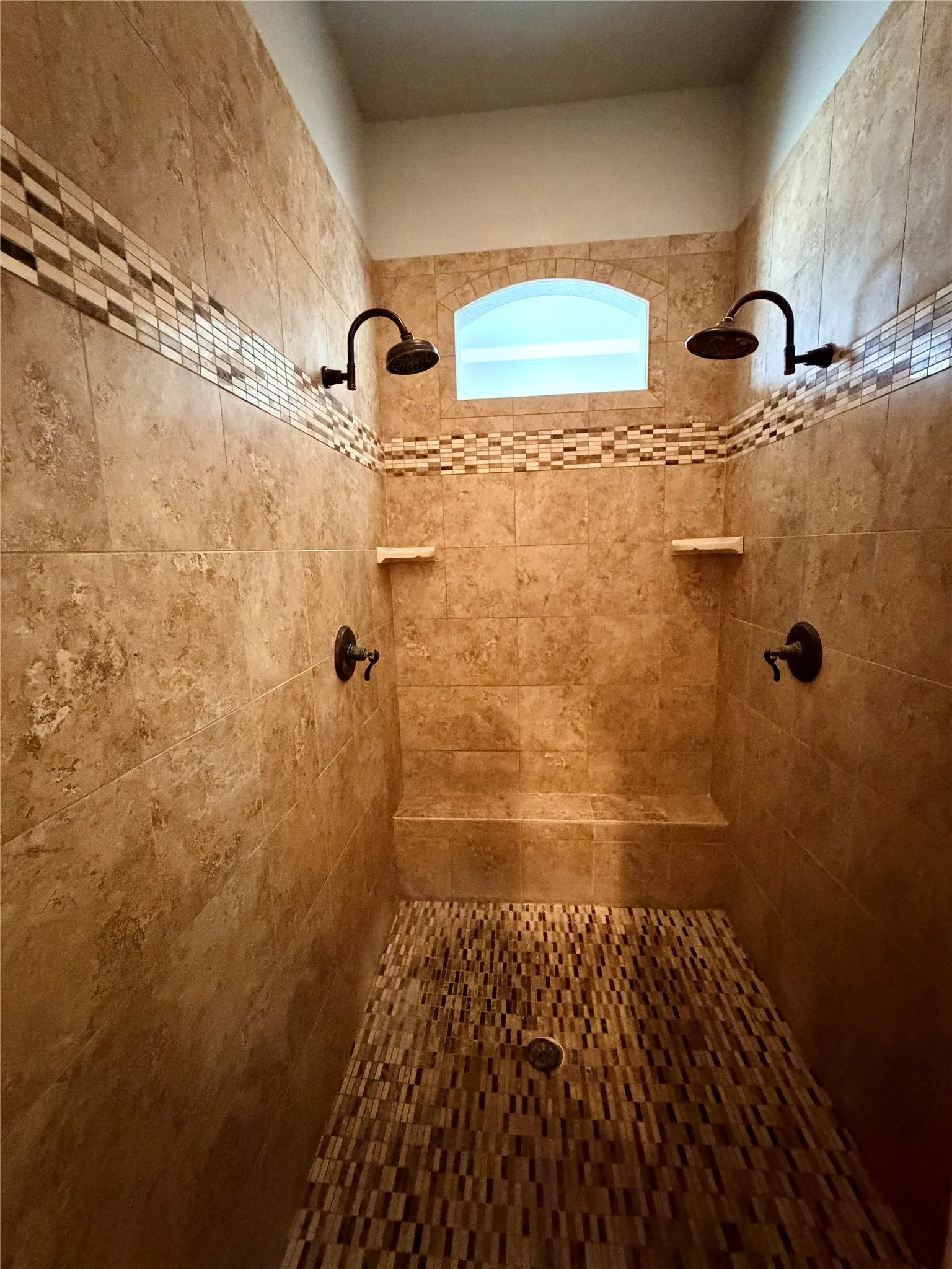 Tiled shower featuring dual showerheads, a tiled bench, an arched transom window, and a mosaic tile floor