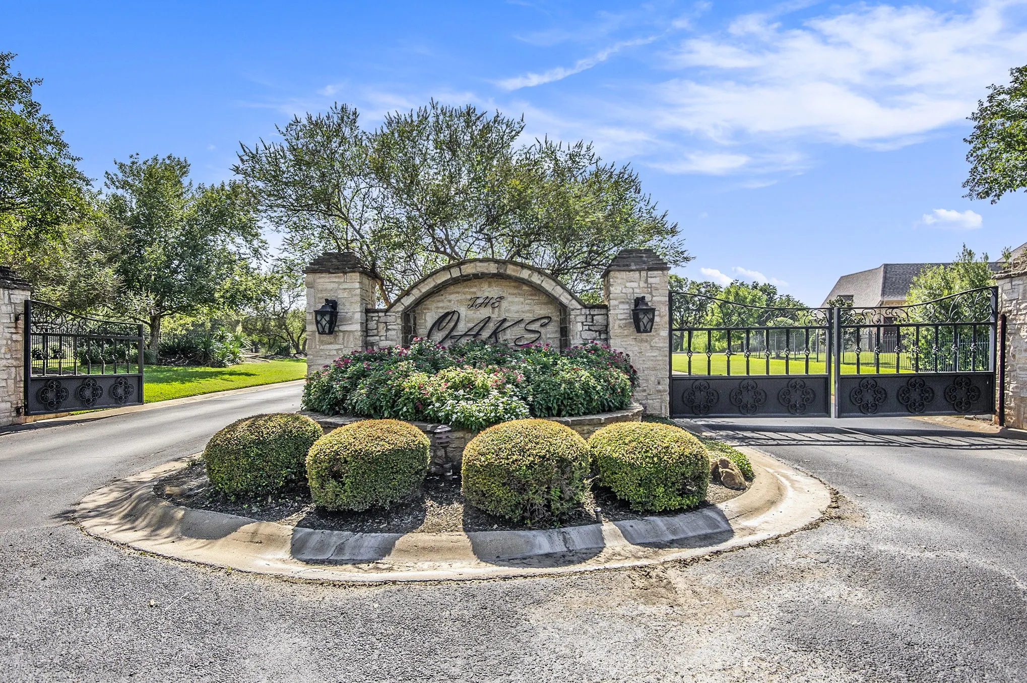 Stone-clad entry columns with decorative wrought iron gates, a landscaped center island, and mature trees