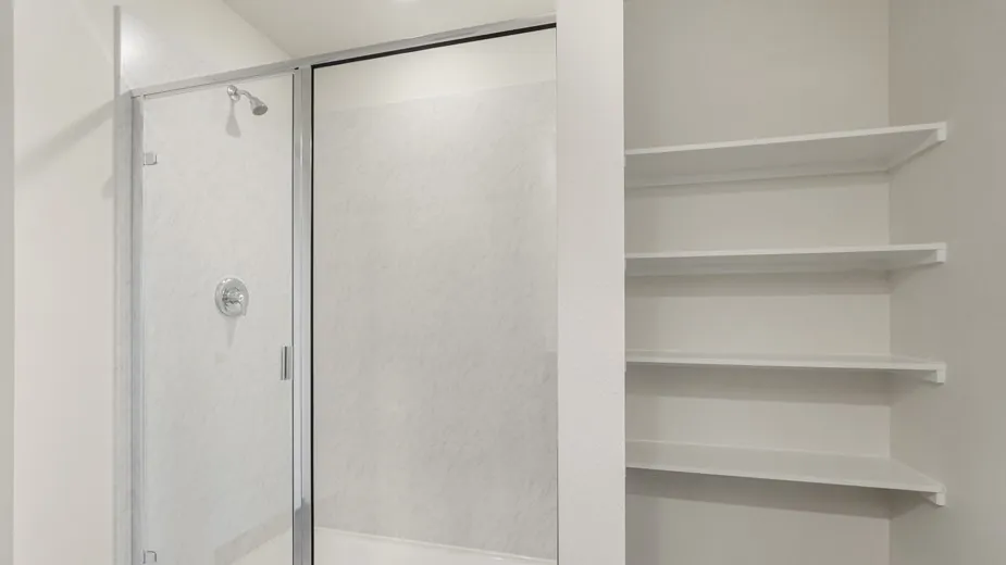 Shower enclosure with clear glass door, chrome showerhead, and circular control, accompanied by built-in shelving