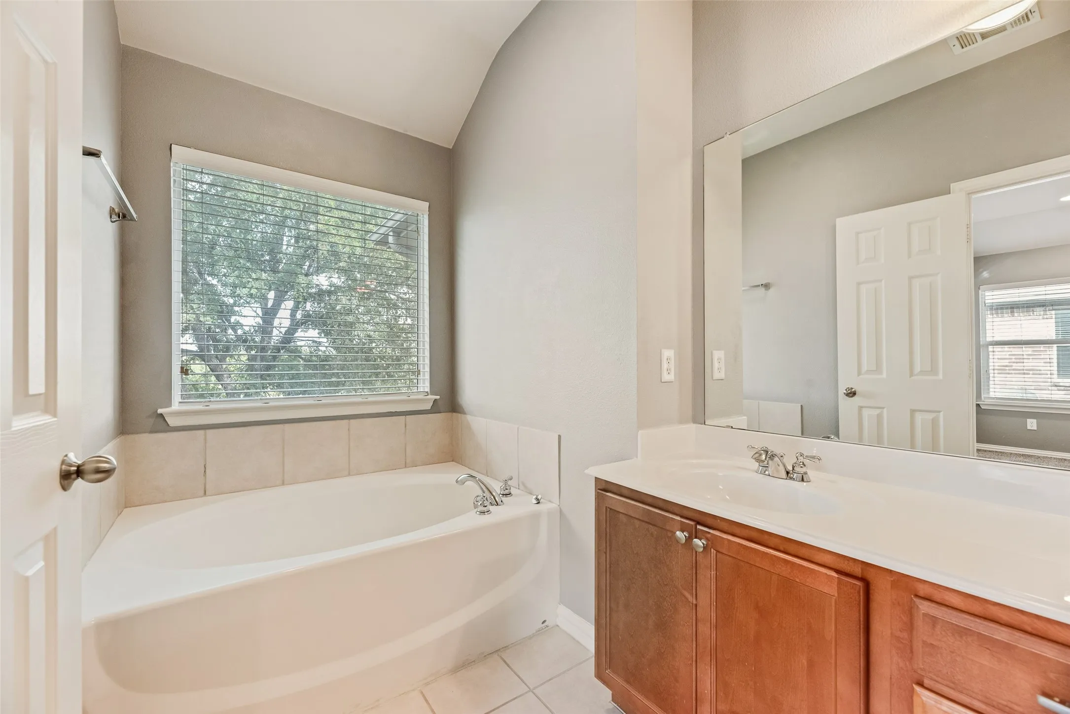 Built-in corner bathtub with tiled surround, single vanity with integrated sink, wood-finish cabinetry, and a large wall-mounted mirror
