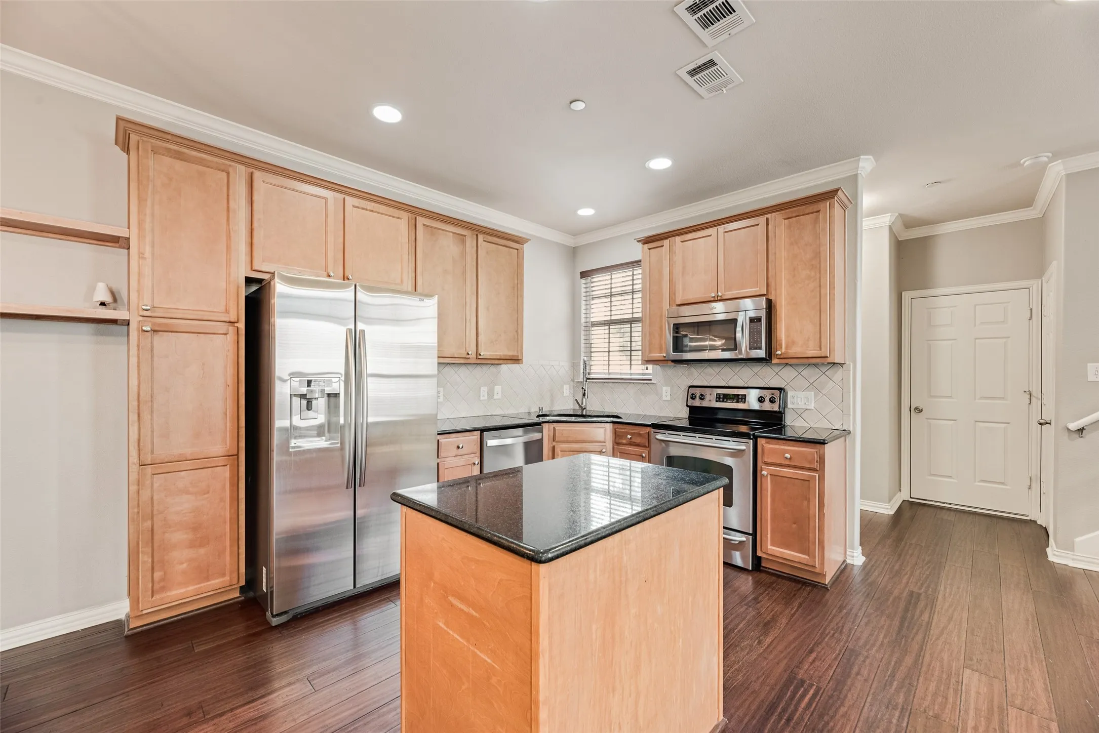 Kitchen featuring wood-finish flooring, stainless steel appliances, dark countertops, a tile backsplash, and light wood cabinetry
