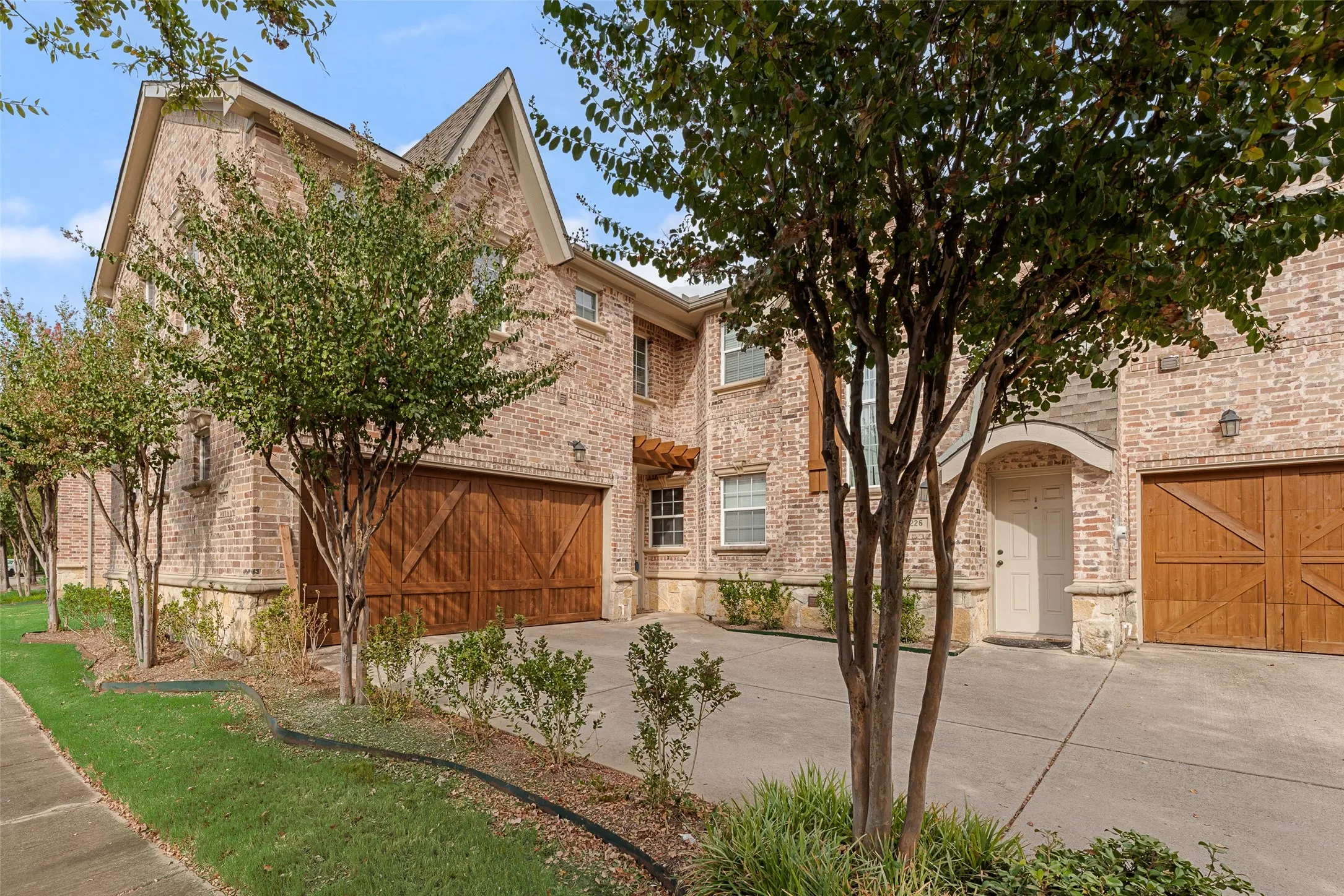 Brick exterior featuring multiple gables, wood-finish garage doors, concrete driveway, and established plantings