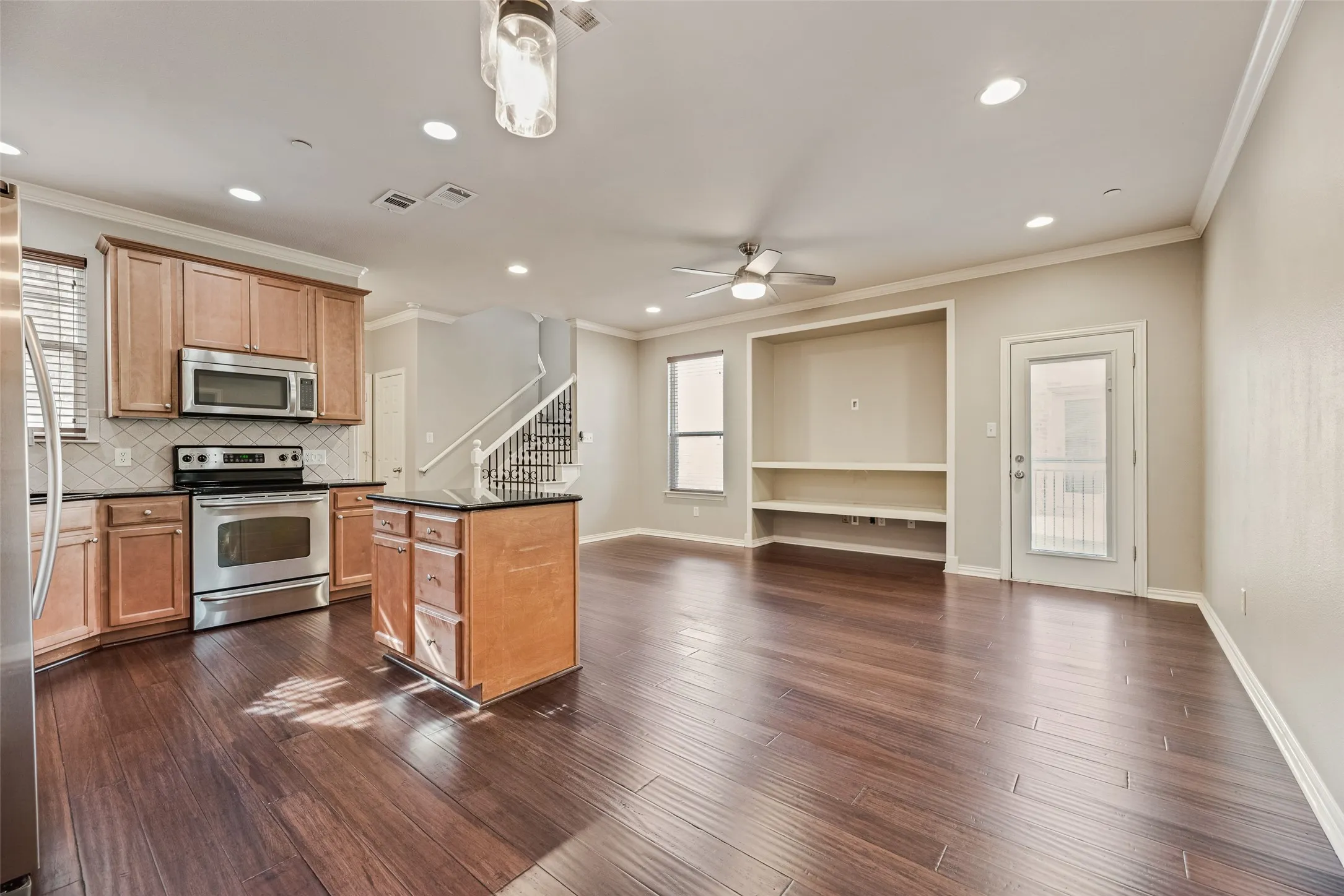 Open concept living space featuring wood-finish flooring, a kitchen with stainless steel appliances, a center island with dark countertops, built-in shelving, and a prominent staircase with metal balusters