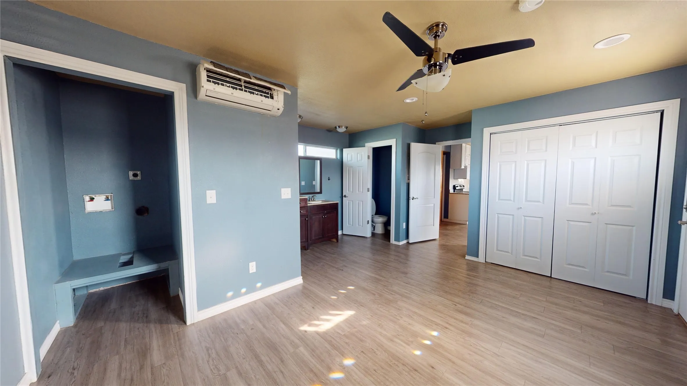 Wood-finish flooring, ceiling fan with integrated lighting, built-in closet with bi-fold doors, wall-mounted air conditioning unit, and a built-in laundry alcove