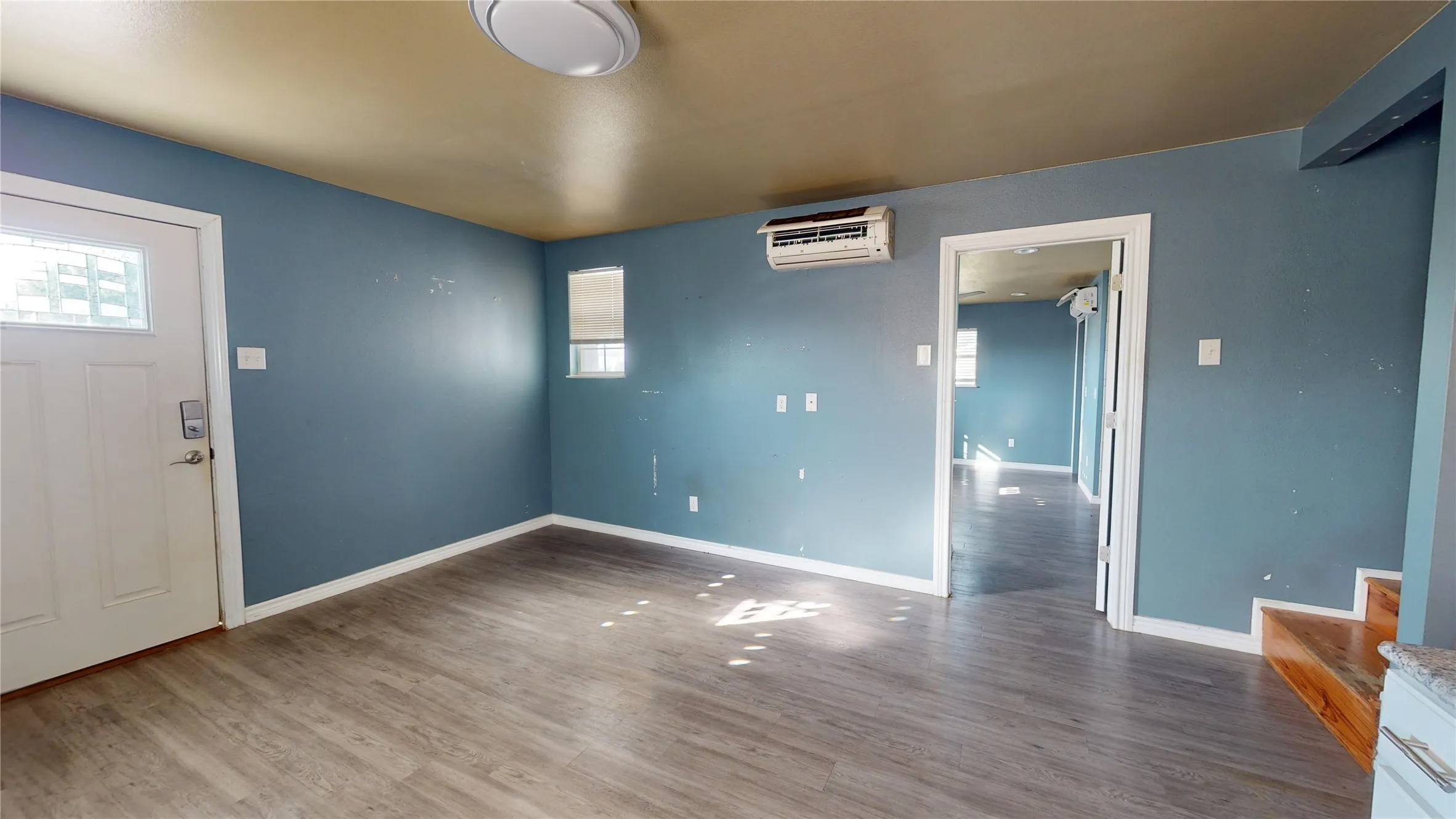 Wood-finish flooring throughout, featuring a white paneled entry door with frosted glass, a window with horizontal blinds, and a wall-mounted climate control unit