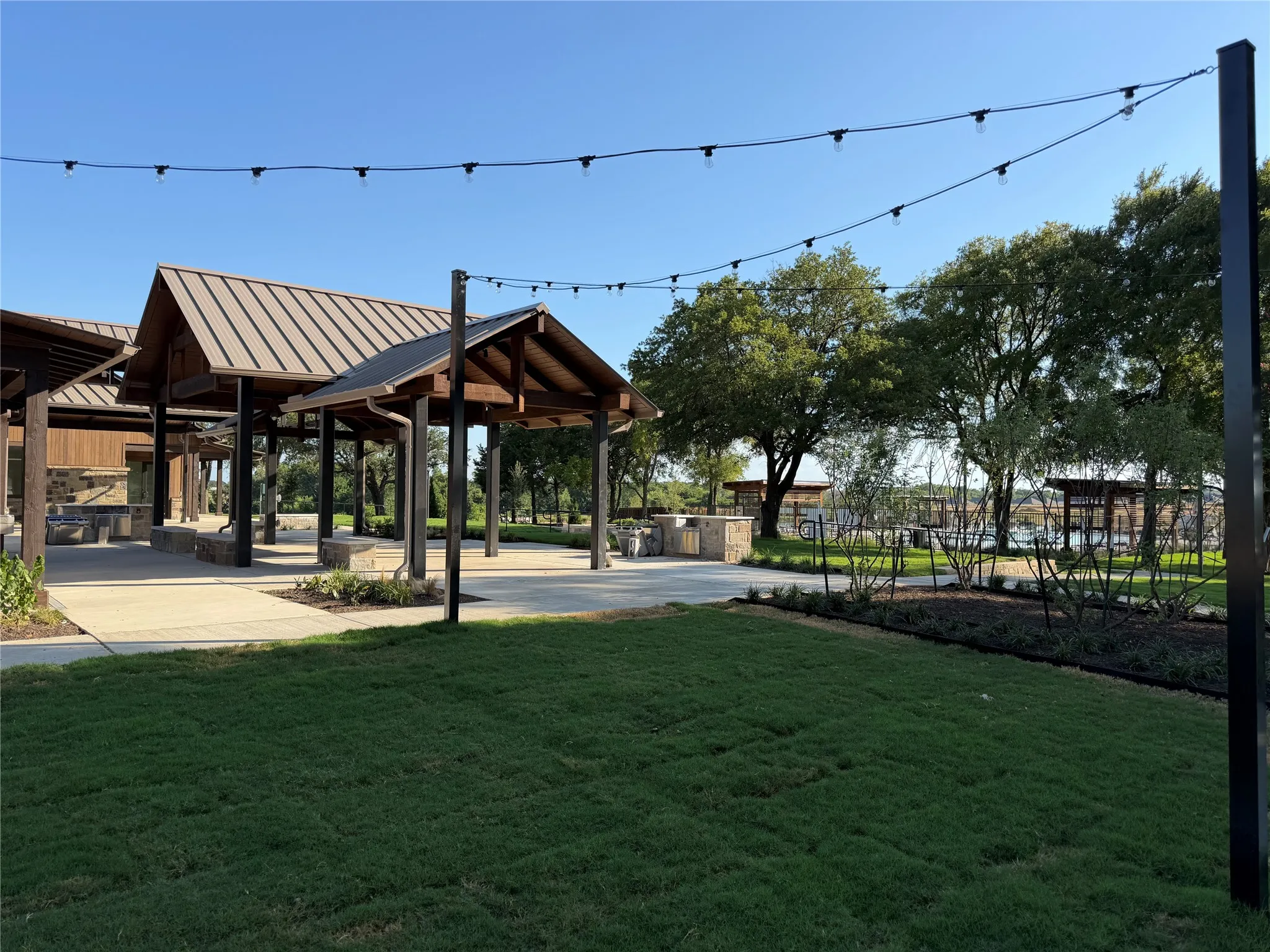 Expansive green space featuring string lighting, multiple covered pavilions with metal roofs, paved pathways, and mature trees