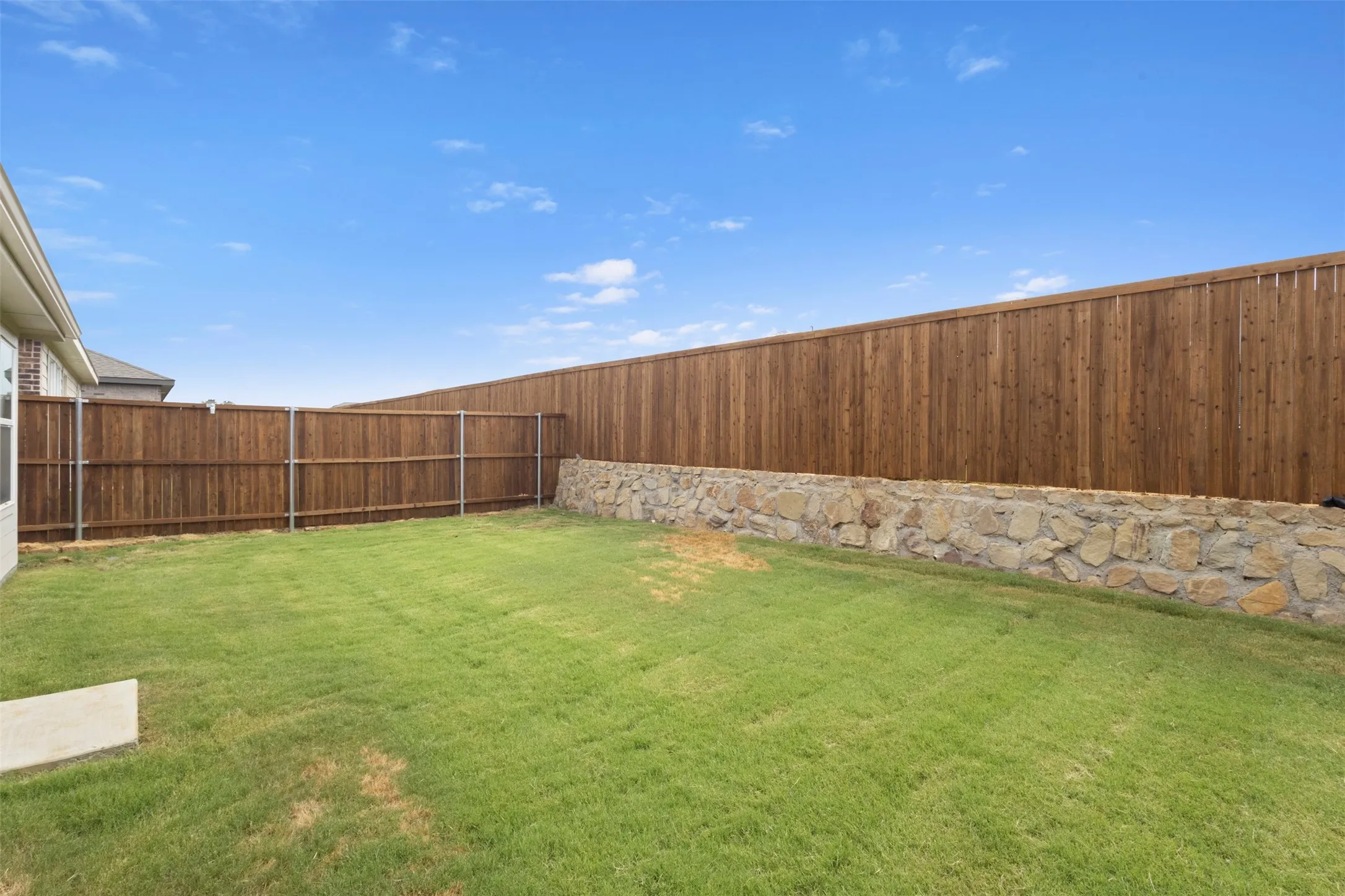 Expansive grassy yard featuring a wood fence, a stone retaining wall, and a concrete patio slab
