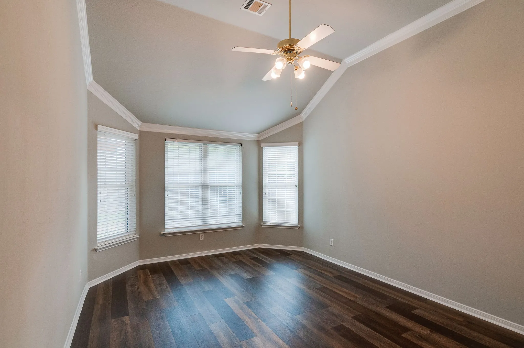 Vaulted ceiling, crown molding, wood-finish flooring, ceiling fan with light fixture, and multiple windows with blinds