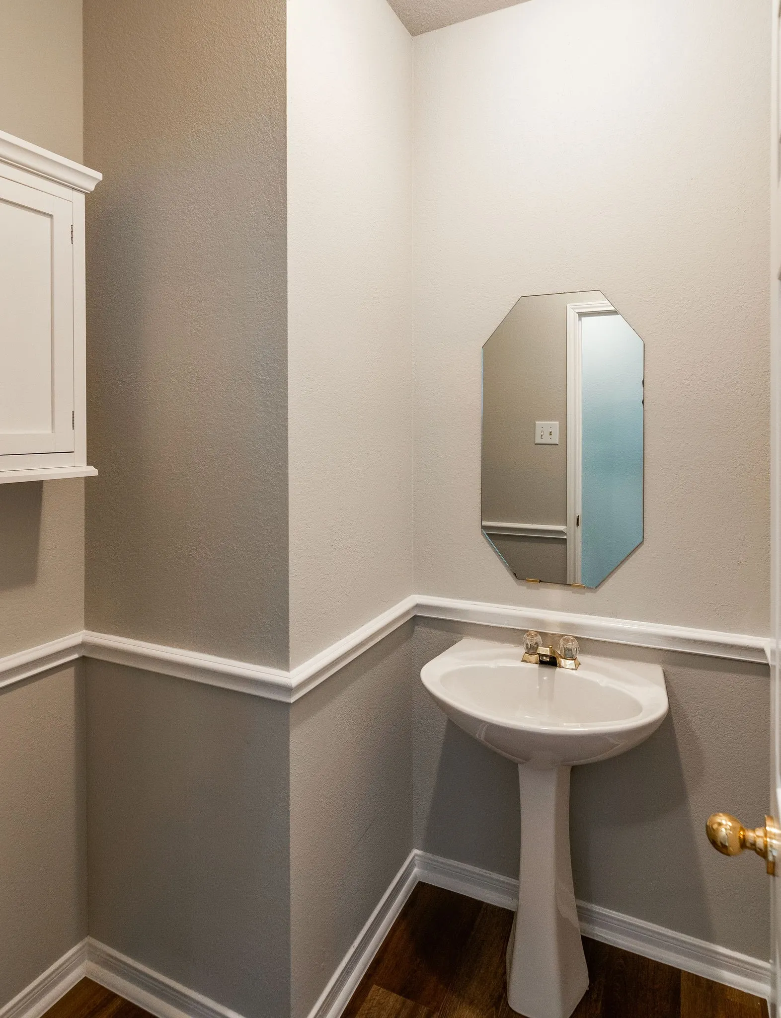 Pedestal sink with brass-finish faucet, octagonal mirror, wood-finish flooring, and white wall cabinetry