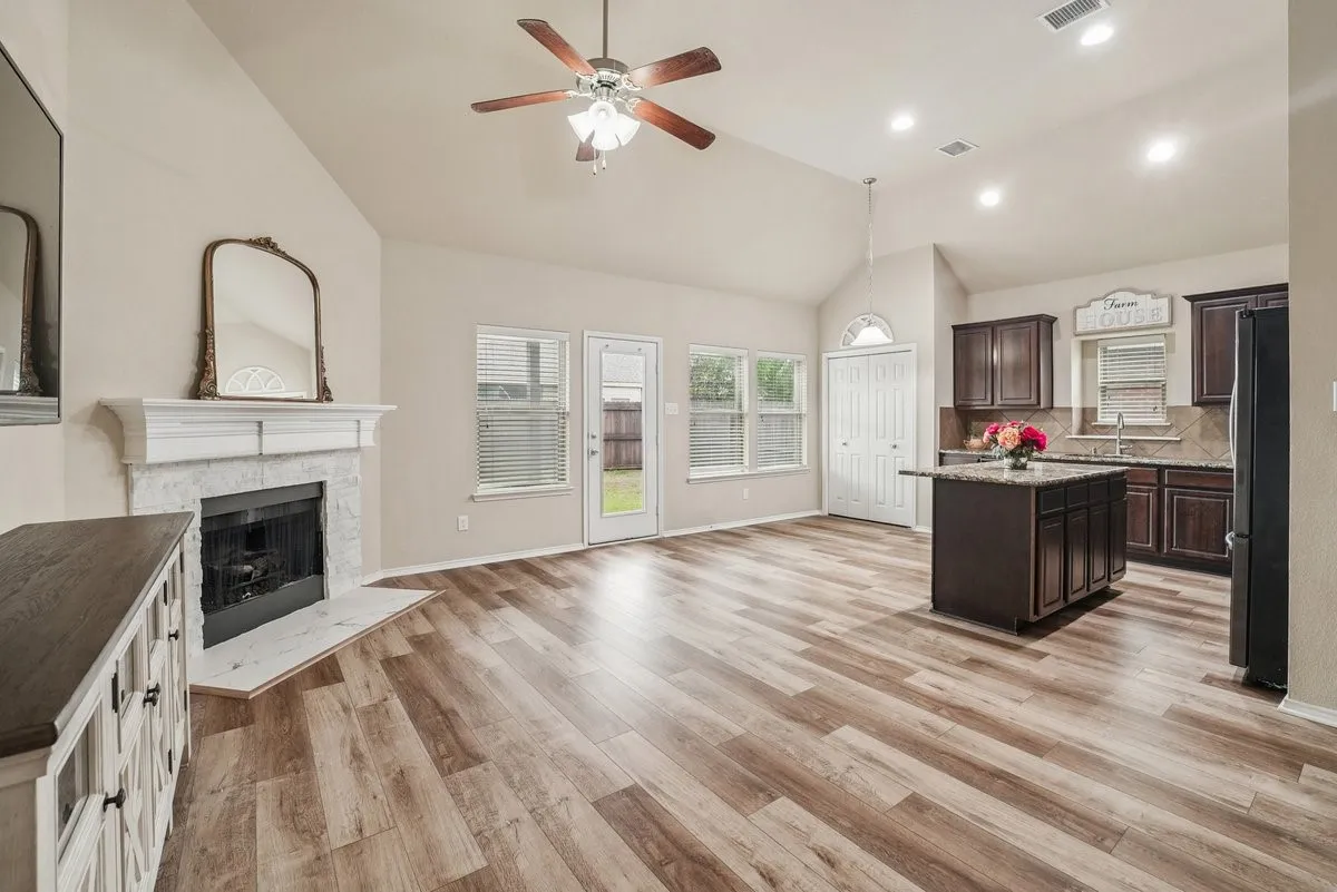 Vaulted ceiling, wood-finish flooring, white mantel fireplace, kitchen island with dark cabinetry, and recessed lighting