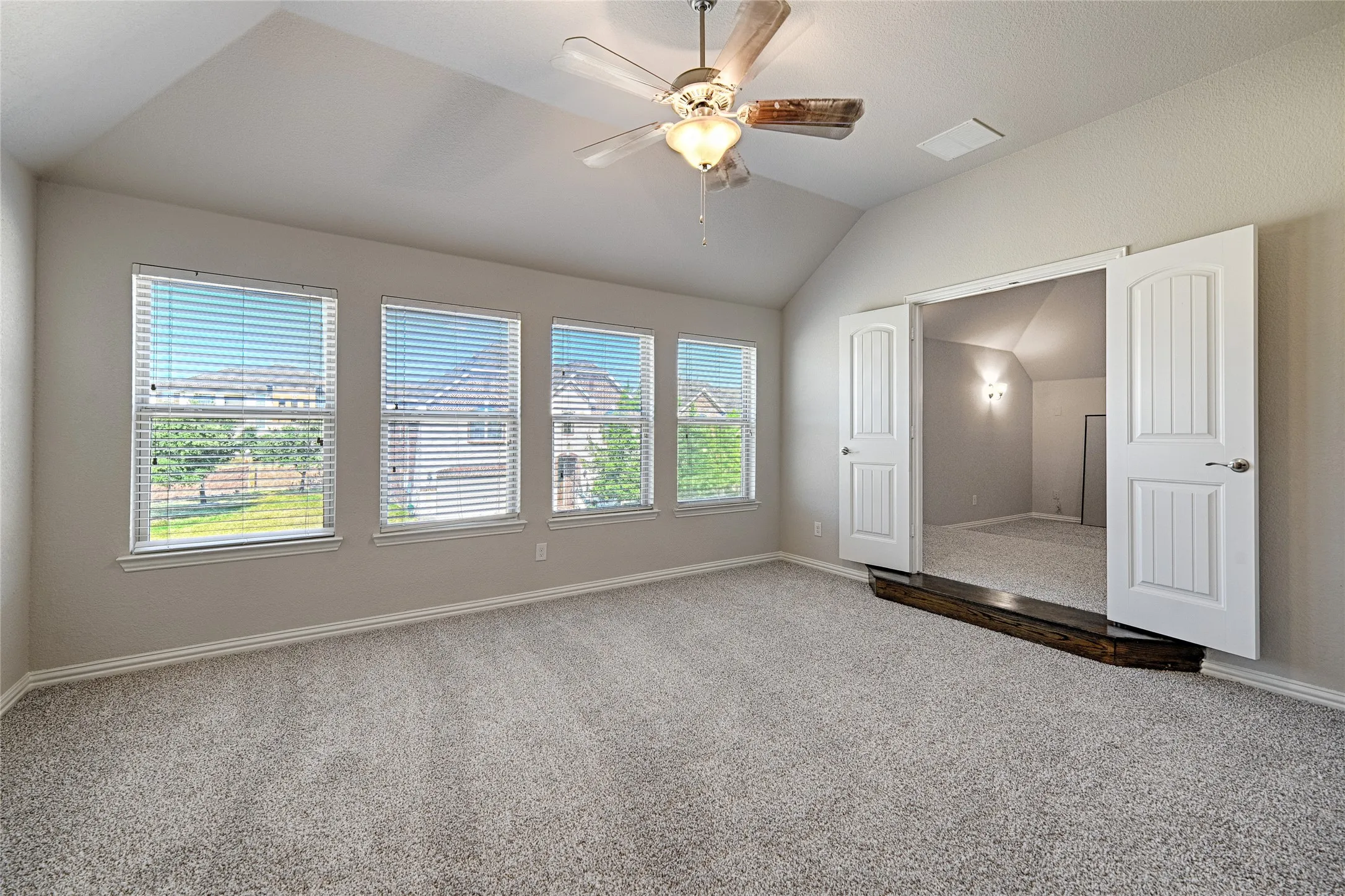 Vaulted ceilings, three large windows, textured carpeting, a ceiling fan with light fixture, and double doors opening to an additional room