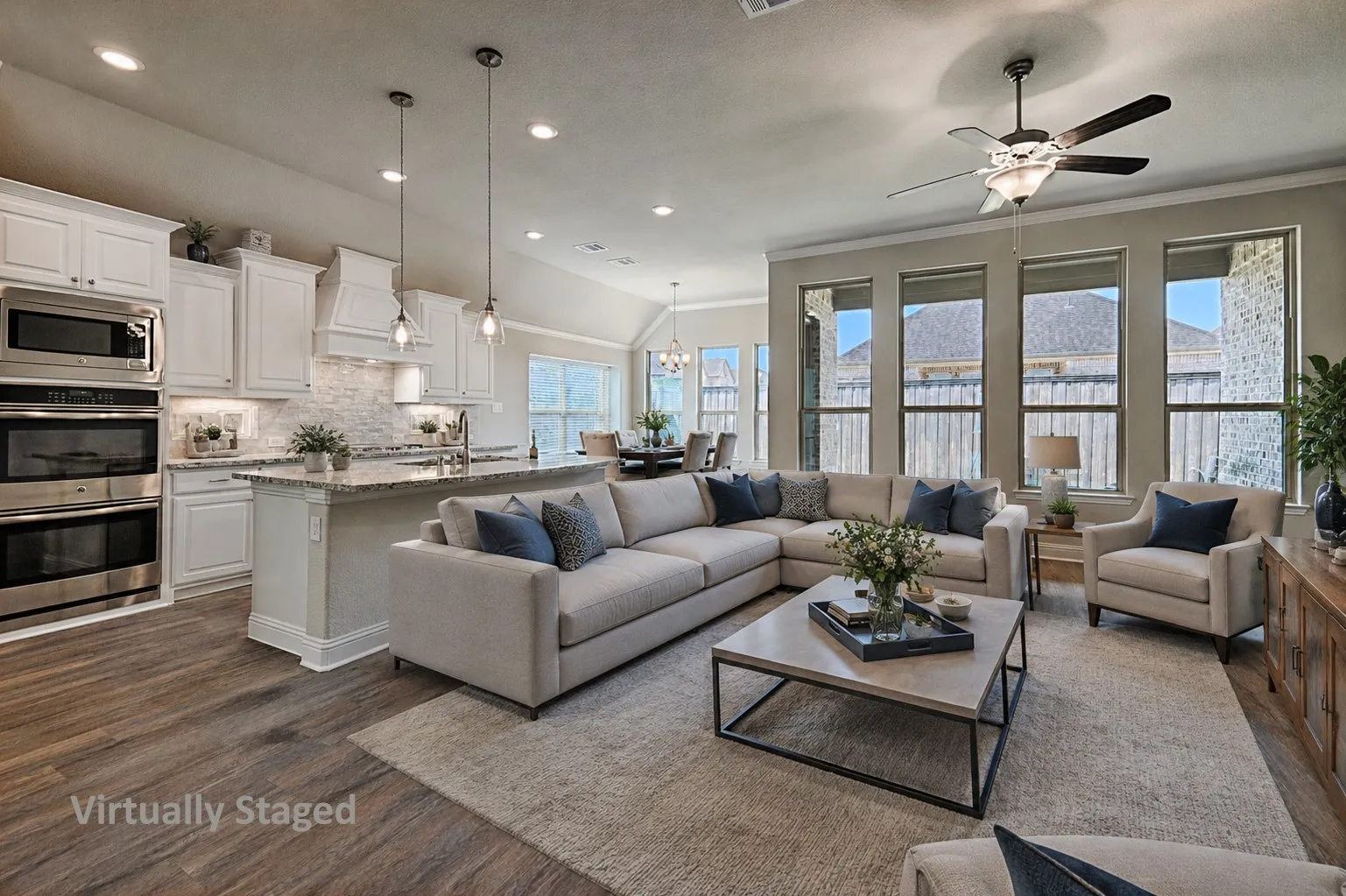 Open-concept living space featuring wood-finish flooring, a kitchen island with stone countertop, stainless steel double ovens, white cabinetry, and multiple windows