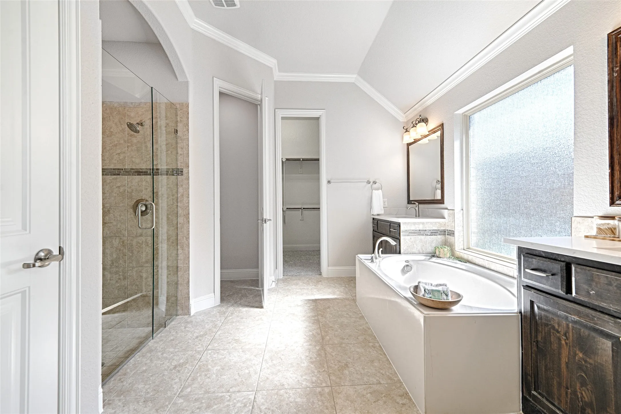 0rimary bathroom. Tiled flooring throughout, walk-in shower with glass enclosure, built-in soaking tub, dual vanities with integrated sinks, and frosted window
