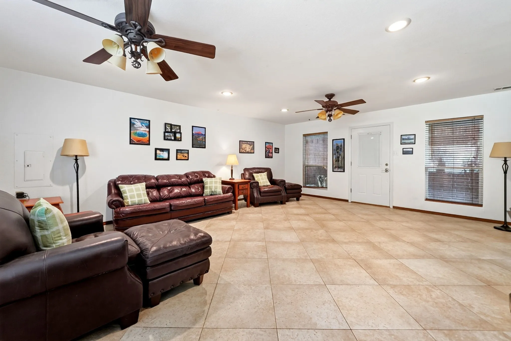 Spacious room featuring light-toned tile flooring, two ceiling fans with integrated lighting, multiple recessed ceiling lights, and two windows with horizontal blinds