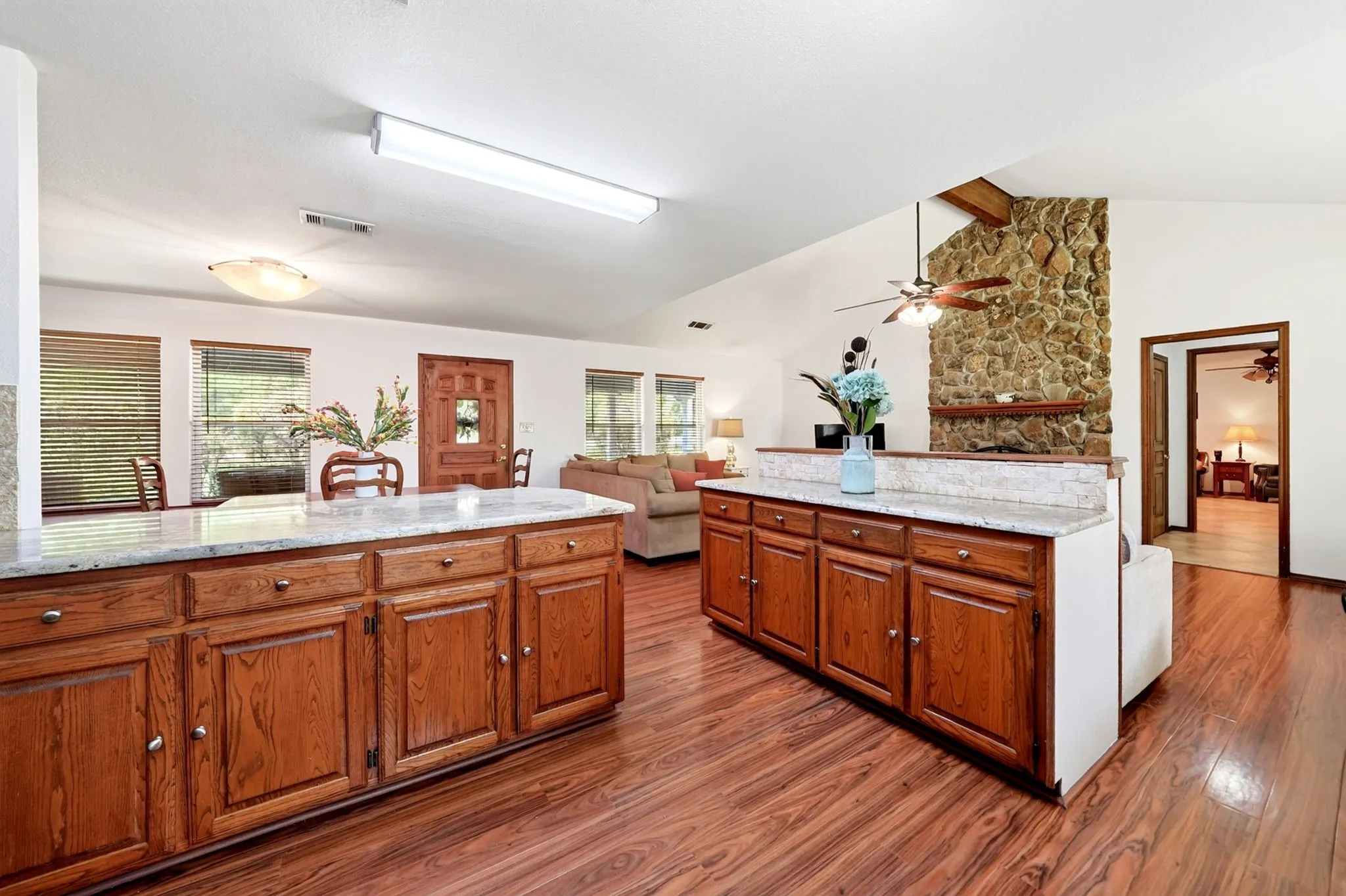 Wood-finish flooring throughout, substantial stone-clad fireplace, wood cabinetry with light countertops, vaulted ceiling with exposed beam, and a ceiling fan