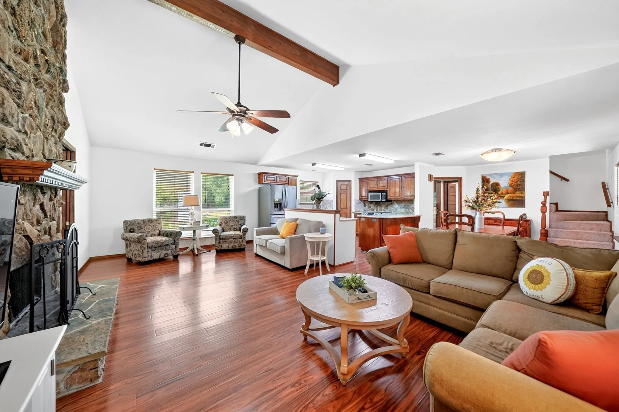 Vaulted ceiling with exposed wood beam, stone-clad fireplace with stone hearth, wood-finish flooring, recessed lighting, and a ceiling fan