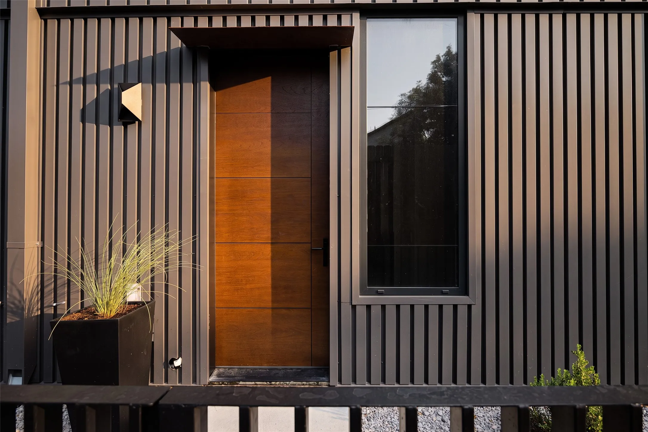 Vertical slat siding, wood-paneled door, rectangular window, exterior wall light, and an overhead door canopy