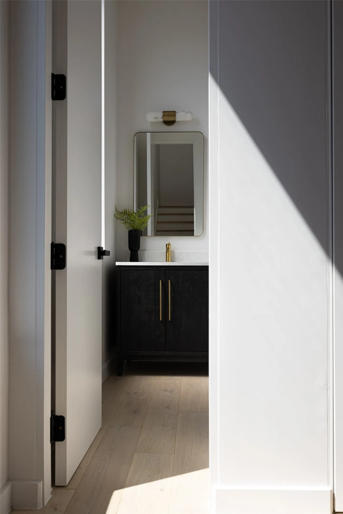 Wood-finish flooring leading to a black vanity with gold hardware, a rectangular mirror, and a wall-mounted light fixture