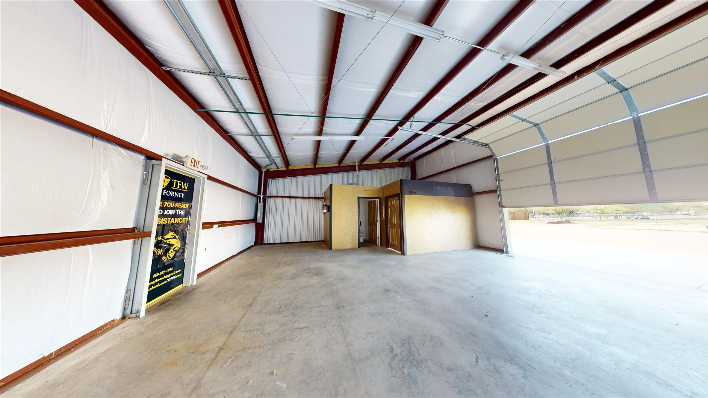 Spacious industrial interior featuring a concrete floor, exposed metal beam ceiling, fluorescent lighting, and a large roll-up door