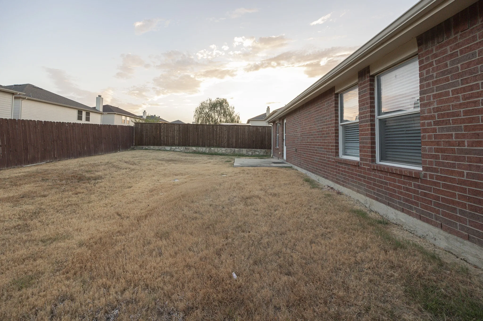 Expansive backyard featuring a wood privacy fence, concrete patio, and brick exterior with multiple windows