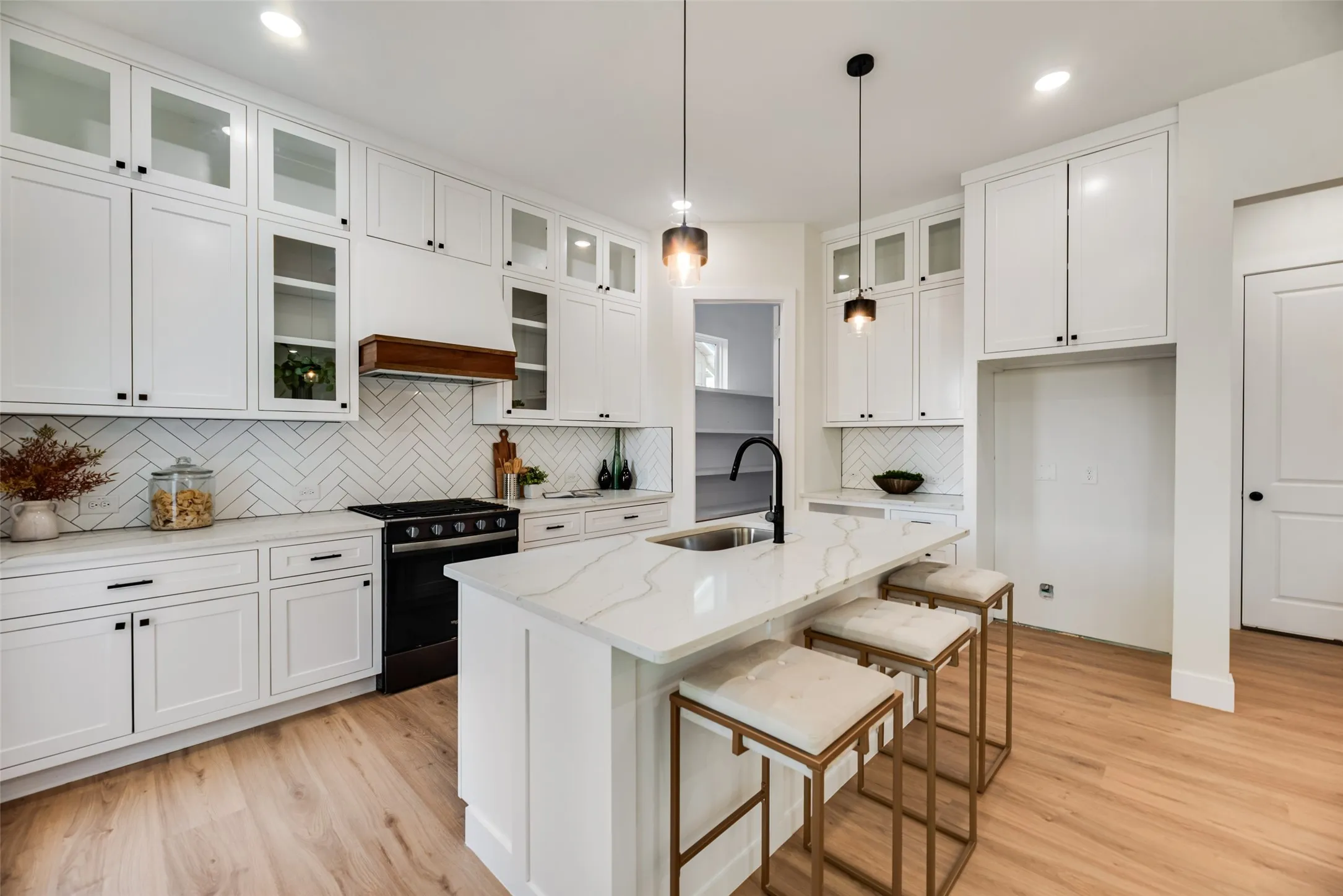 White cabinetry with glass-front uppers, stainless-steel sink with a matte black faucet, herringbone tile backsplash, light wood-finish flooring, and white countertops