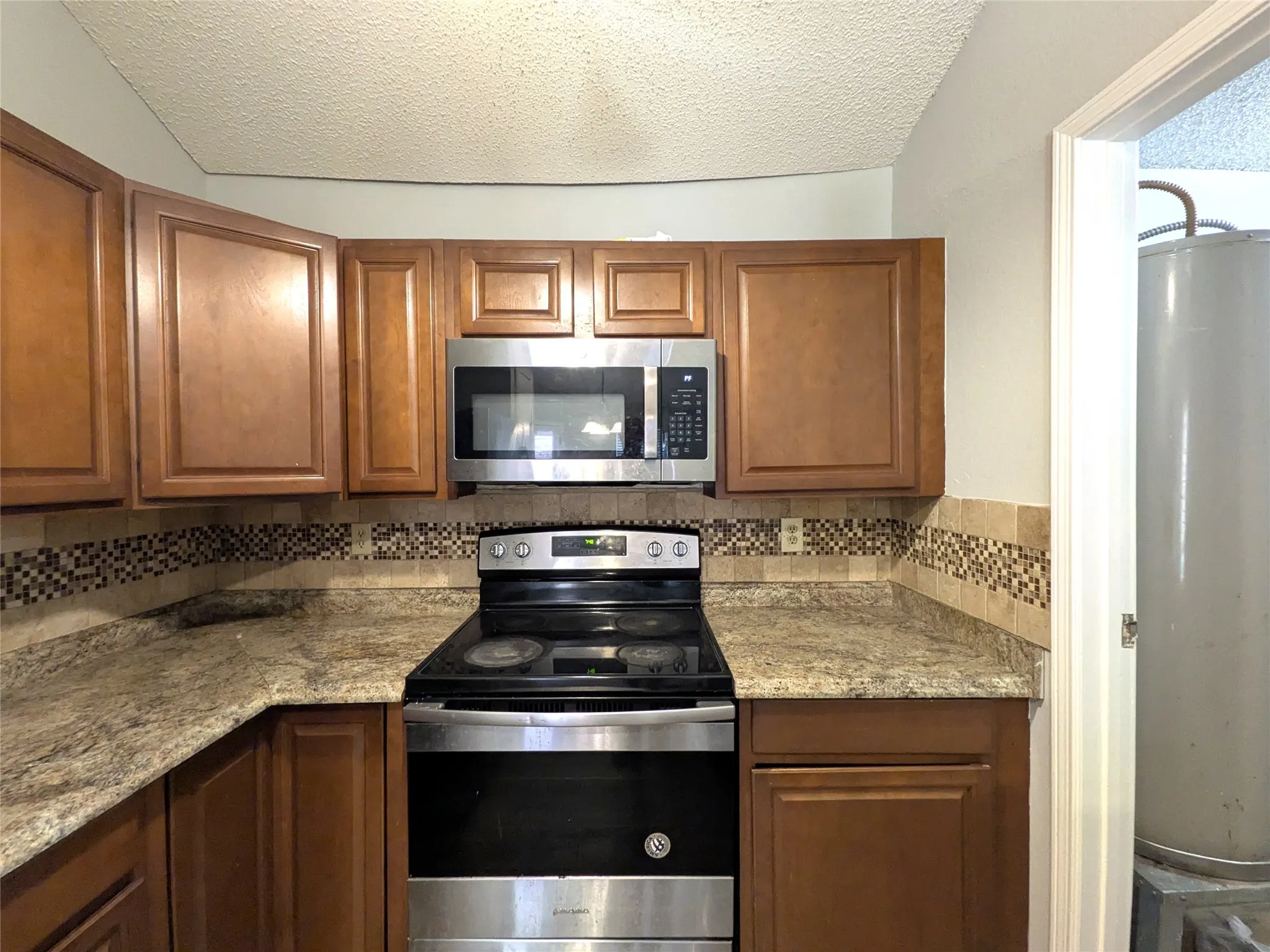 Wood-finish cabinetry, stainless steel microwave, stainless steel range, patterned tile backsplash, and light-toned countertops