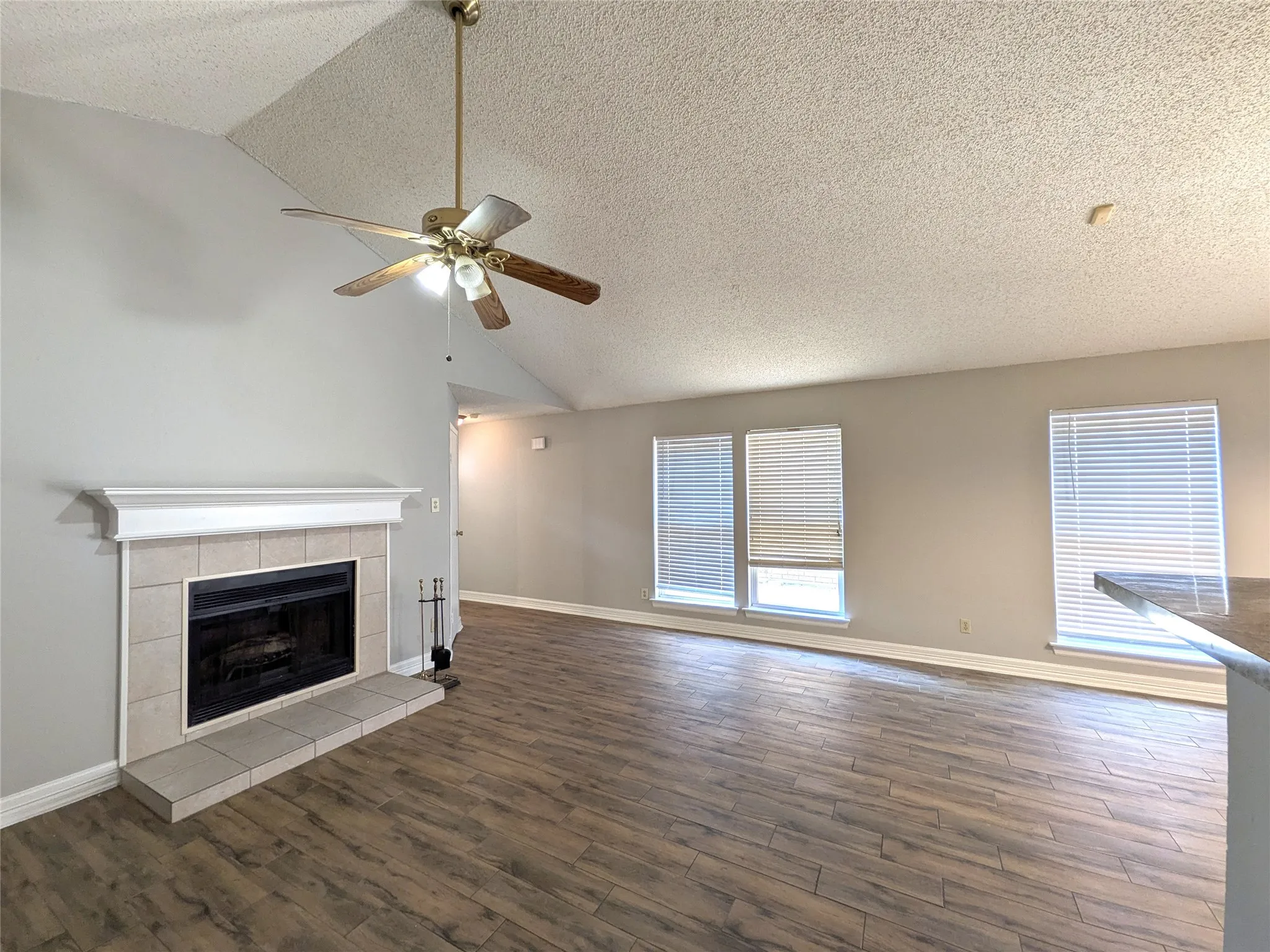 Vaulted ceiling, wood-finish flooring, tiled fireplace with mantel, ceiling fan with light fixture, and multiple windows