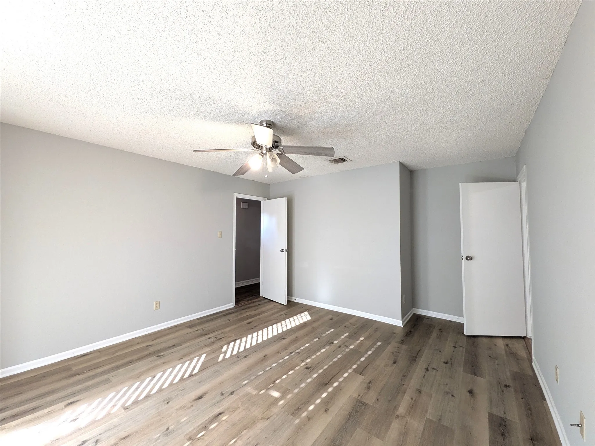 Spacious room featuring wood-finish flooring, light grey painted walls, a textured ceiling, and an overhead ceiling fan with integrated lighting