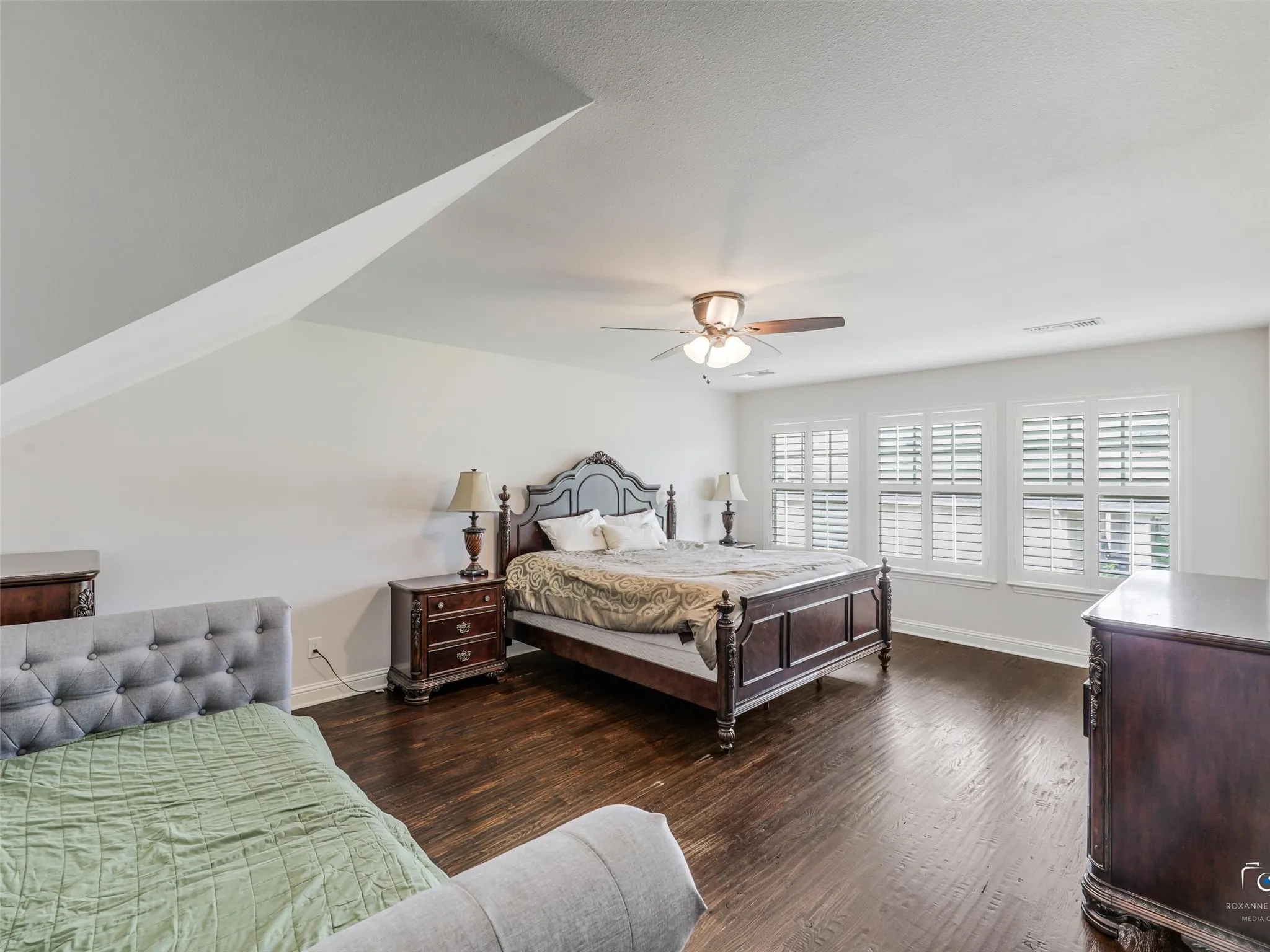 Wood-finish flooring, multiple windows with plantation shutters, ceiling fan with integrated lighting, and sloped ceiling accents