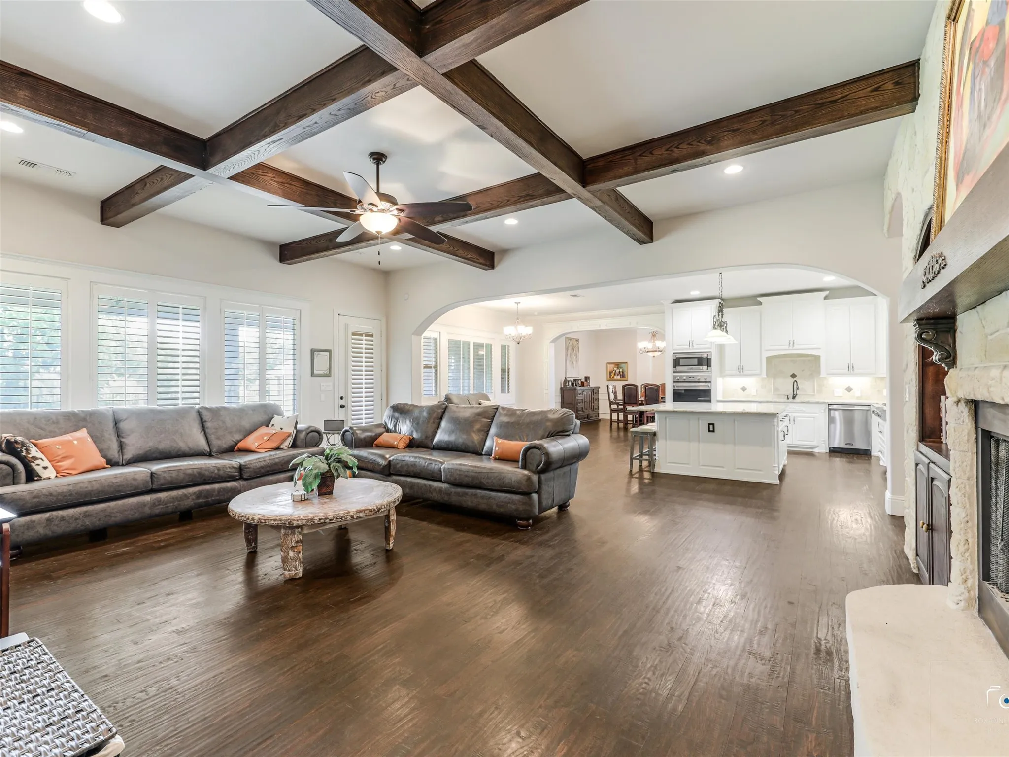 Coffered ceiling with wood beams, dark wood-finish flooring, white plantation shutters, arched kitchen entryway, and a built-in stone fireplace with hearth