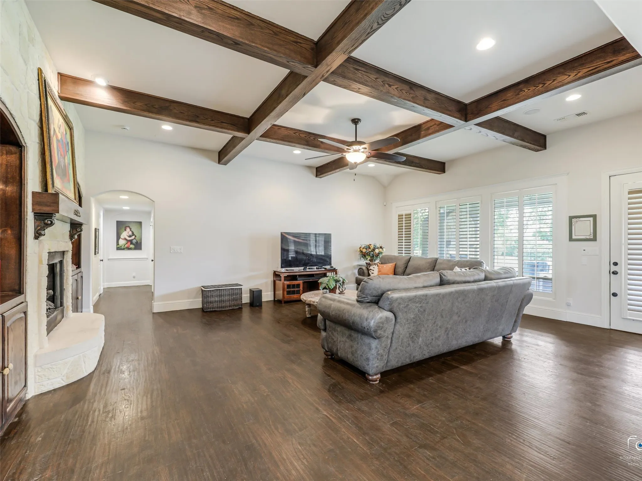Vaulted ceiling with exposed wood beams, recessed lighting, wood-finish flooring, stone-clad fireplace, and multiple windows with plantation shutters