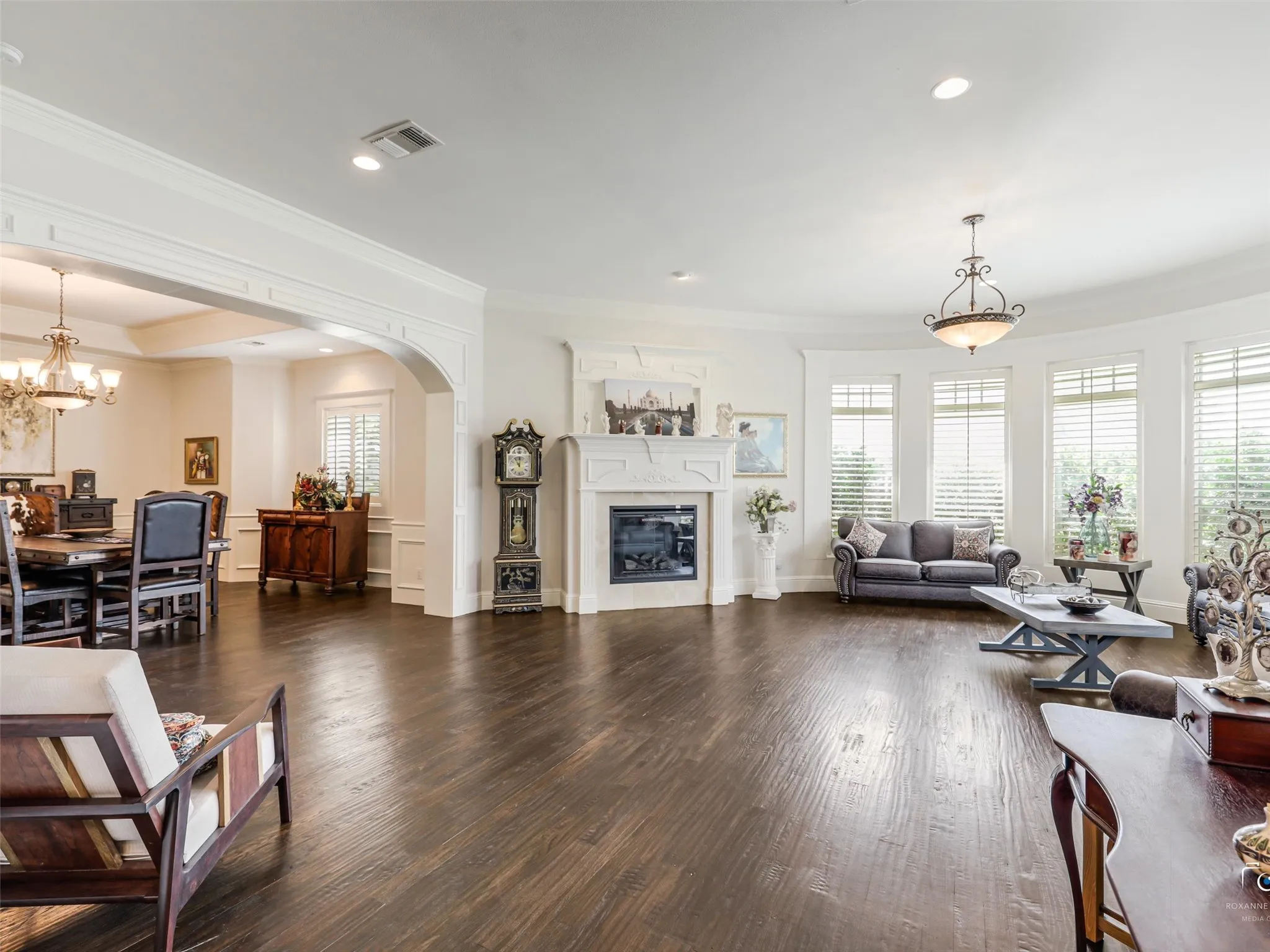Rich wood-finish flooring, a white mantel fireplace, and multiple tall windows with plantation shutters