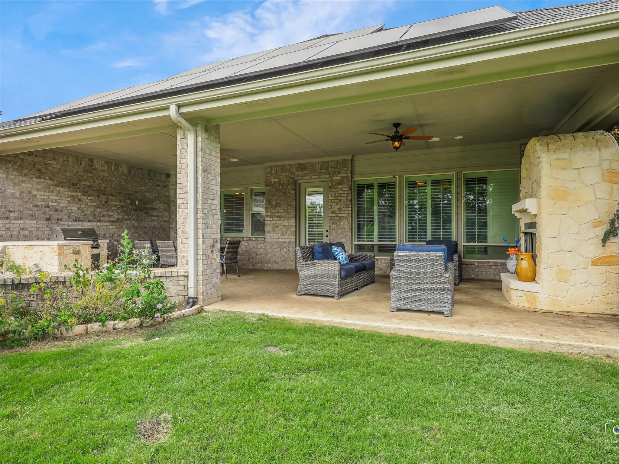 Covered patio featuring a stone-clad outdoor fireplace, an integrated outdoor kitchen with a built-in grill, brick exterior walls, and solar panels on the roof