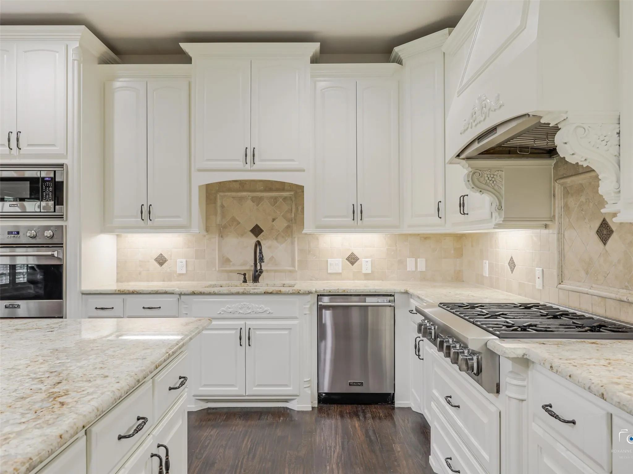 White cabinetry with decorative corbels, stone countertops, stainless steel appliances, tile backsplash, and dark wood-finish flooring