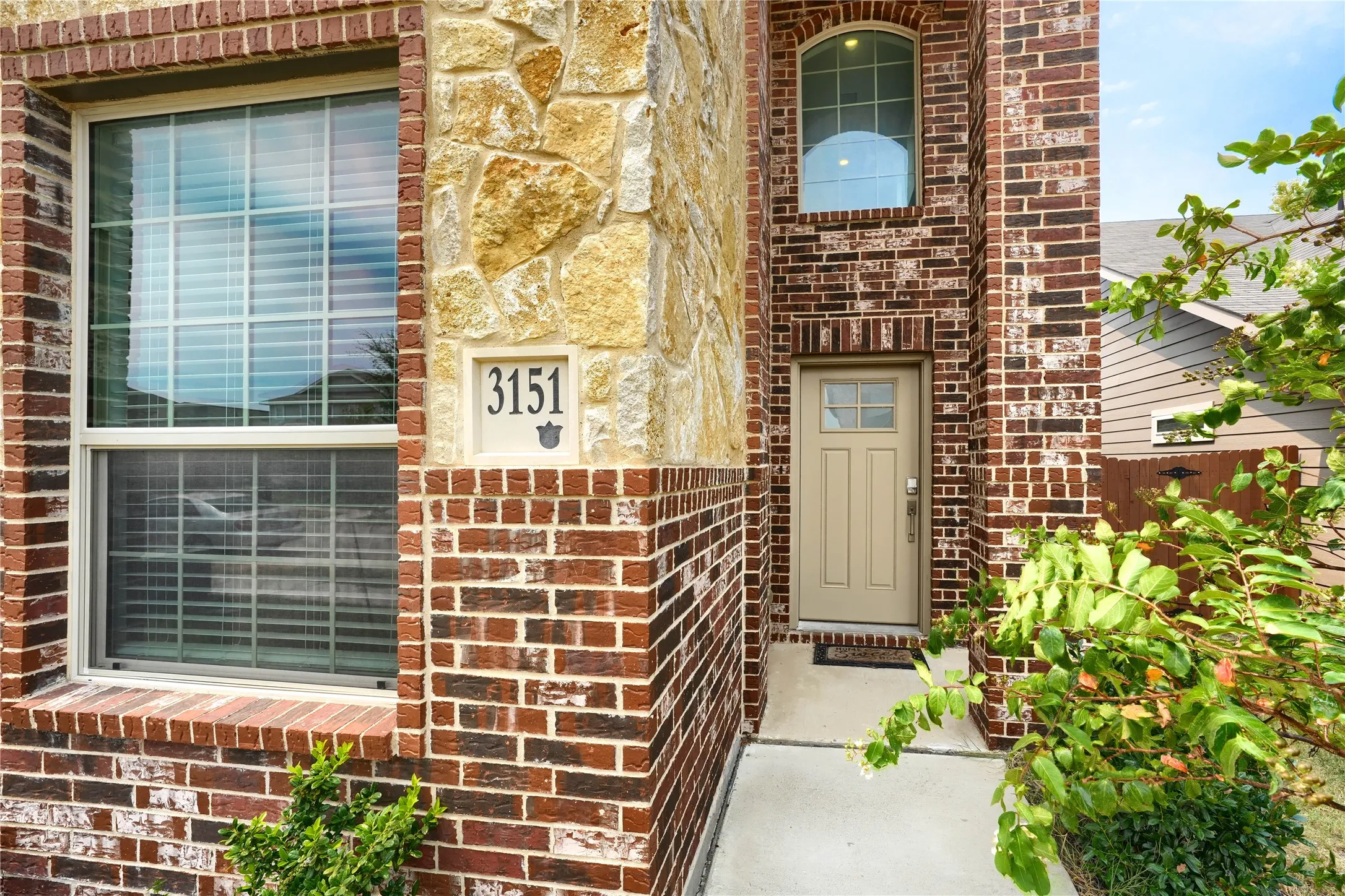 Charming brick and stone exterior featuring a recessed entry with arched transom window, horizontal slider windows with integrated blinds, and lush green landscaping flanking the facade