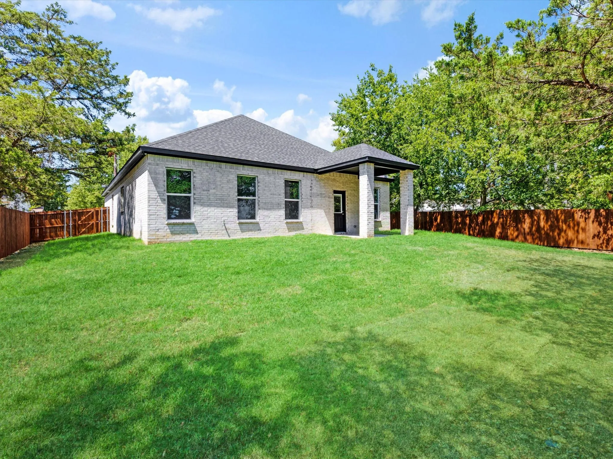 Expansive grassy yard with mature perimeter trees, light brick exterior, dark-framed windows, and a covered patio