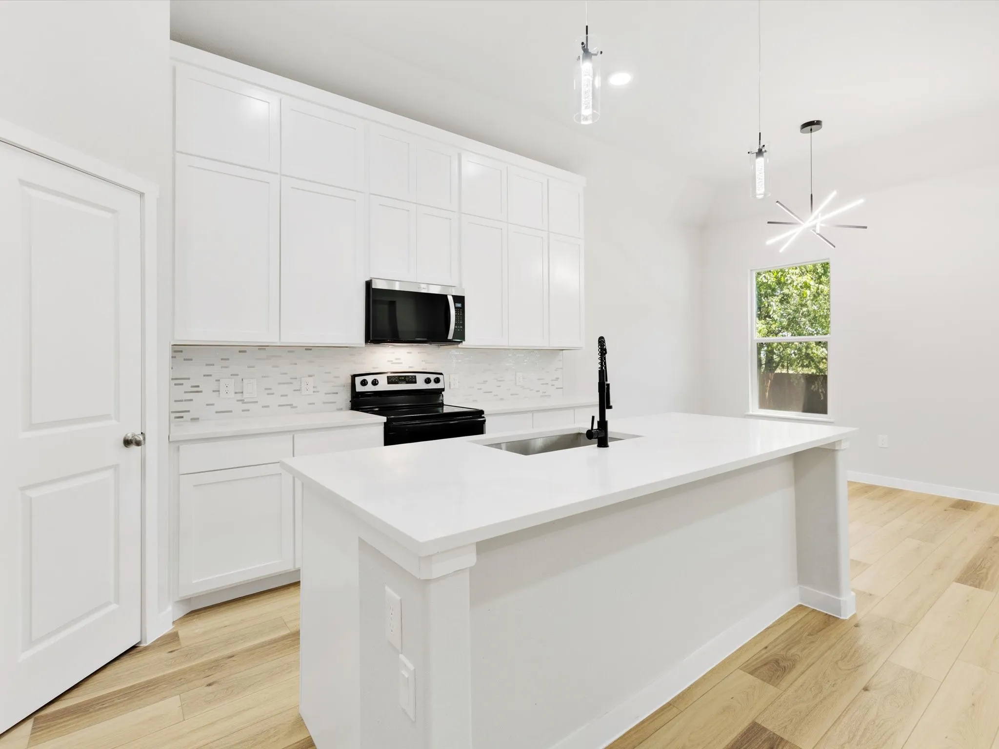 White cabinetry, white countertop island, black appliances, light wood-finish flooring, and contemporary pendant lighting fixtures