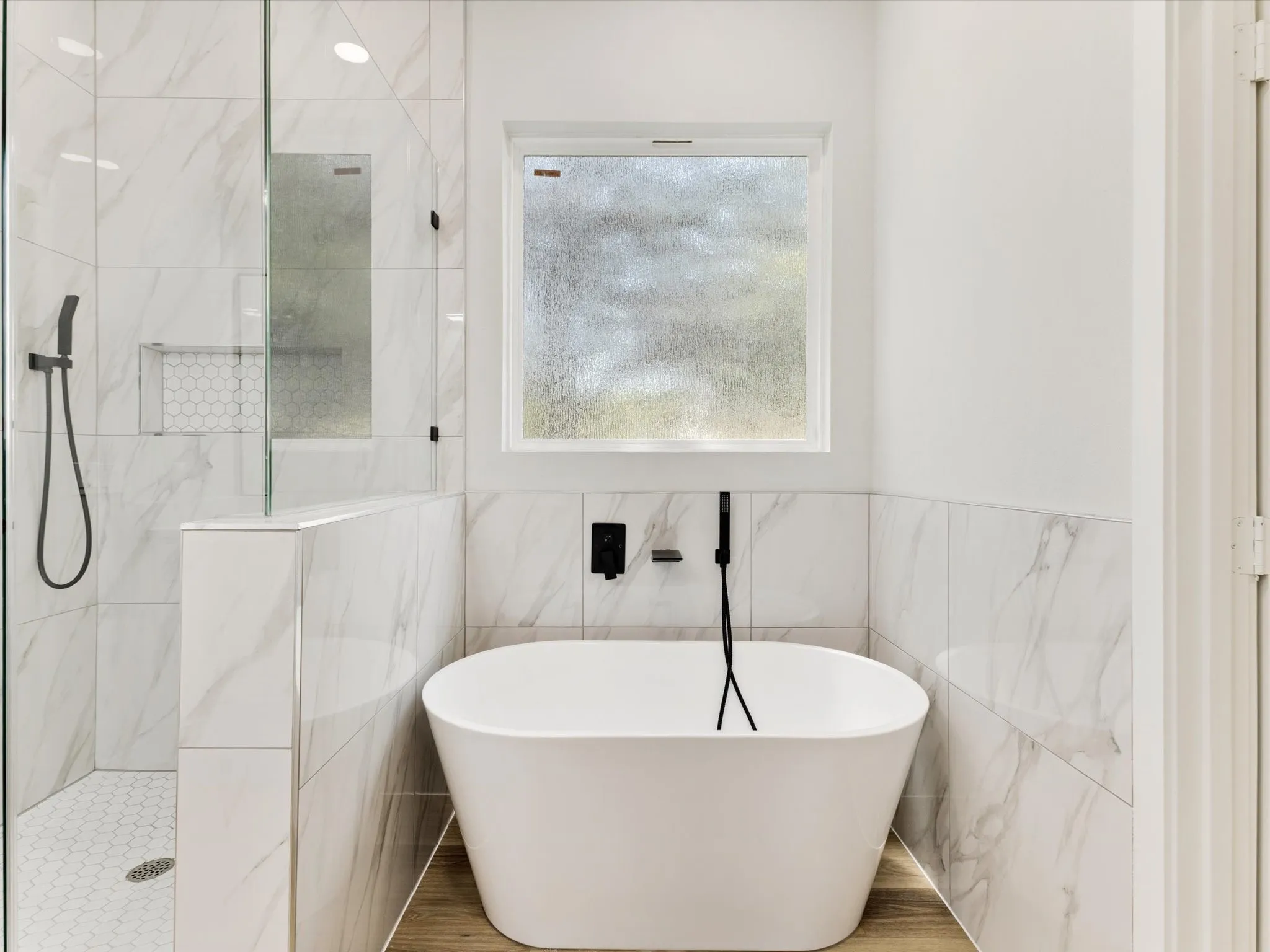 Freestanding soaking tub with matte black fixtures, glass-enclosed shower with hexagon mosaic tile, and large format wall tile