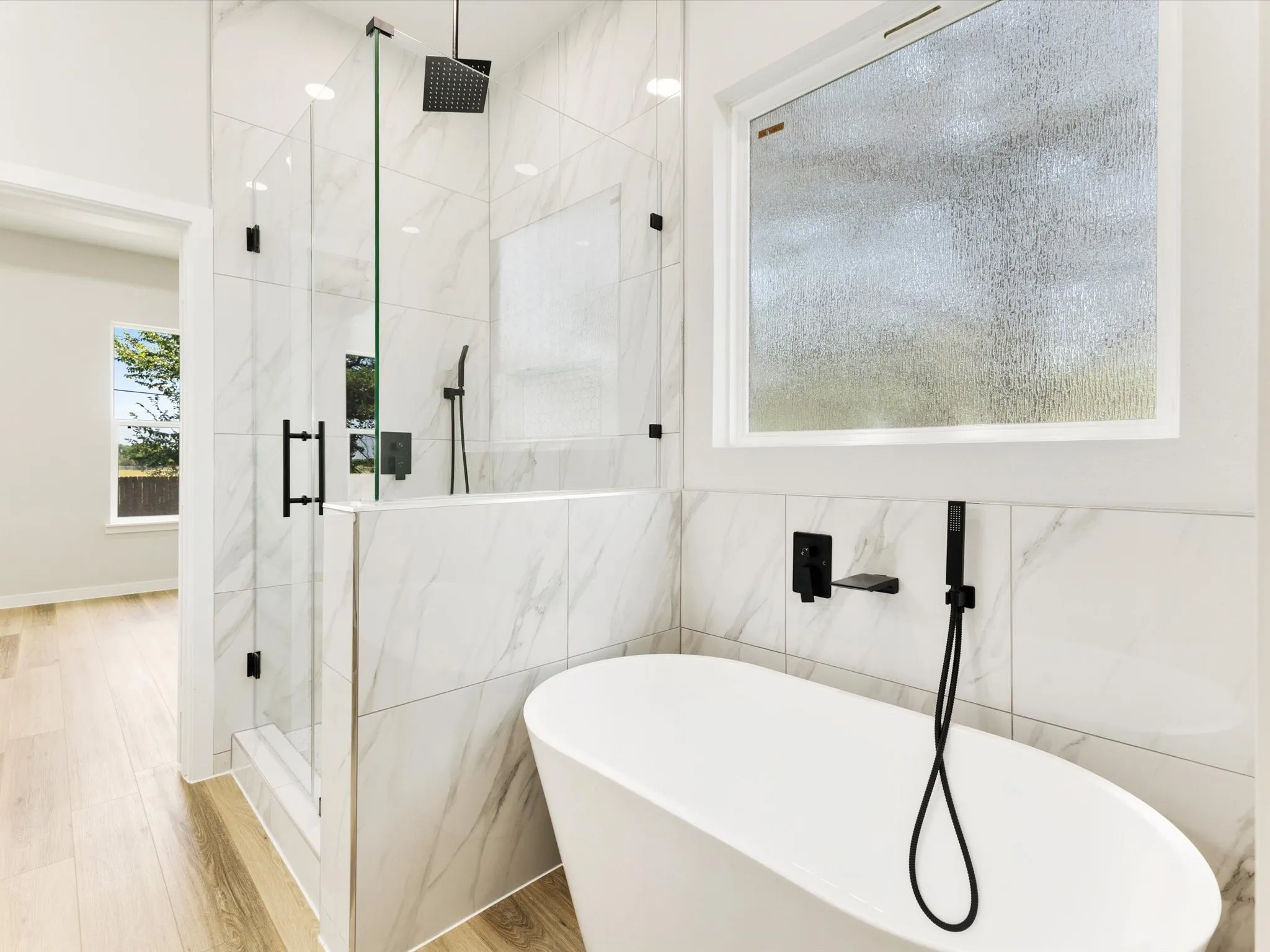 Freestanding soaking tub with matte black fixtures, glass-enclosed shower with rainfall showerhead, large format marble-look tile, and wood-finish flooring