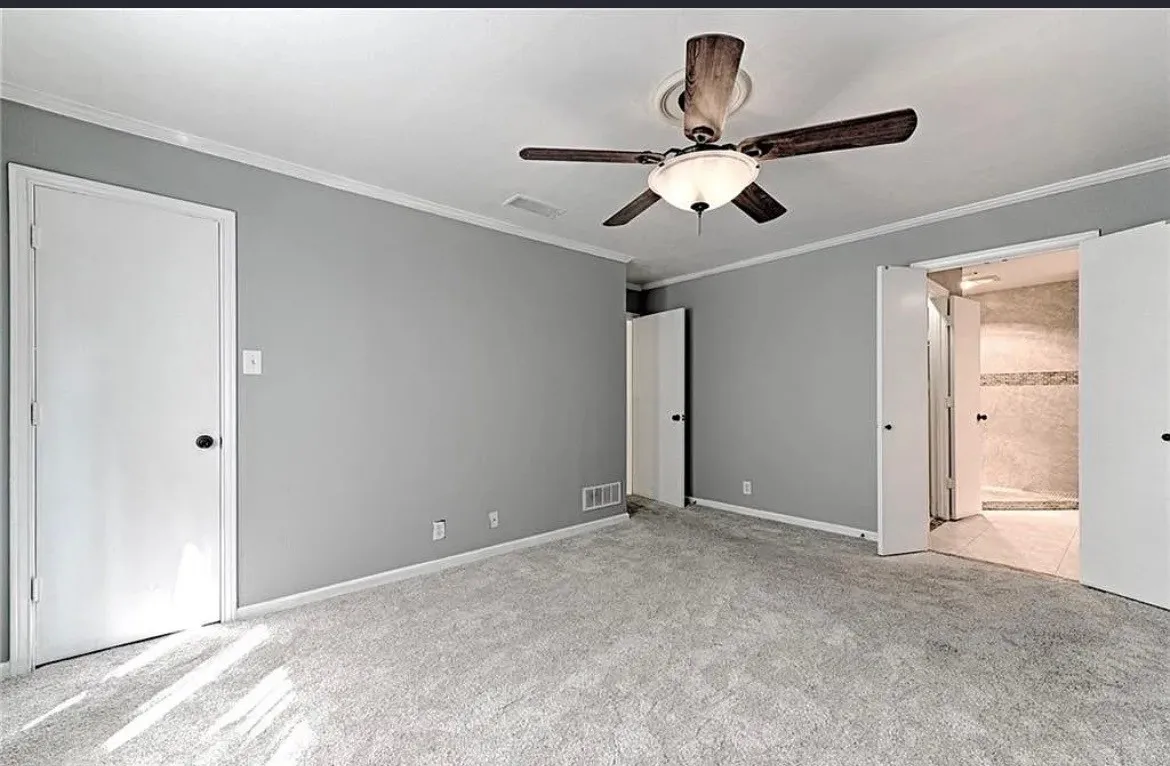 Spacious primary bedroom with neutral gray walls, neutral carpet flooring, ceiling fan with integrated lighting, and direct access to an ensuite bathroom
