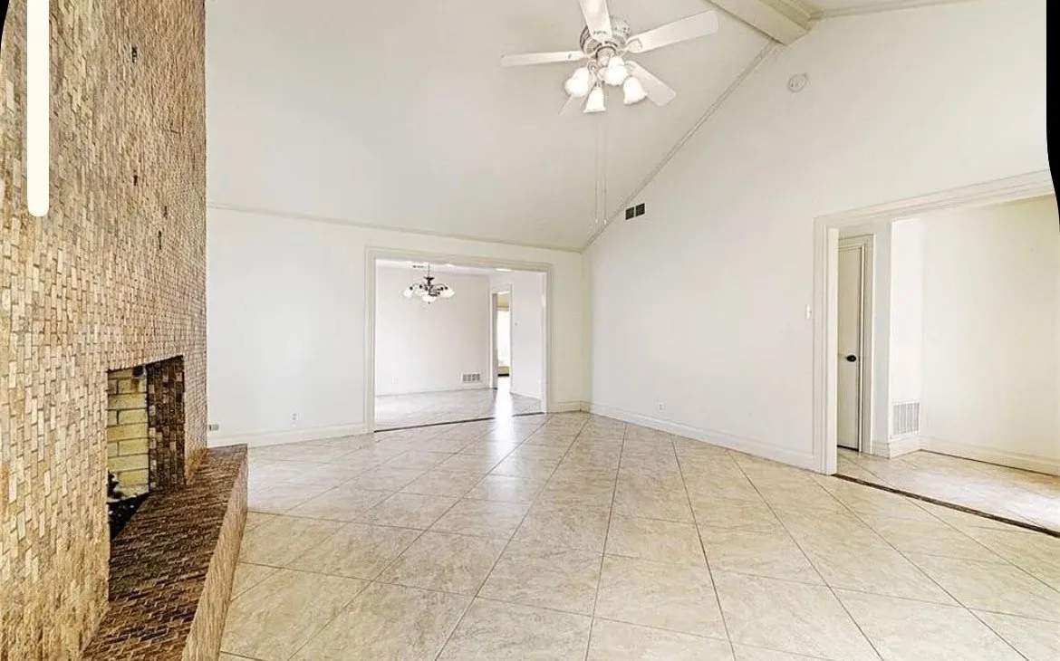Spacious open-concept room featuring neutral tile flooring, vaulted ceiling with ceiling fan, exposed brick chimney with fireplace insert, and multiple entry points