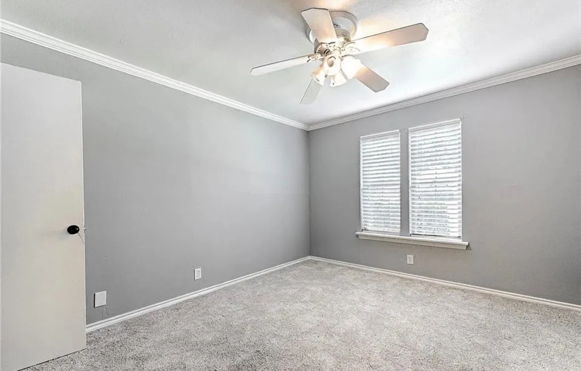 Spacious bedroom with neutral gray walls, neutral carpet flooring, and abundant natural light from dual shuttered windows