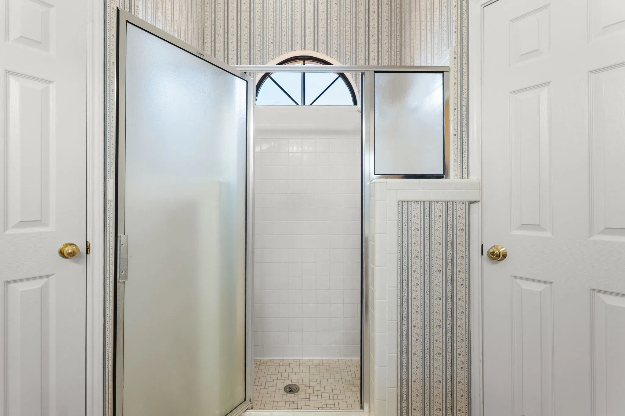 Shower enclosure with frosted glass door, white subway tile surround, tile mosaic shower pan, and arched transom window