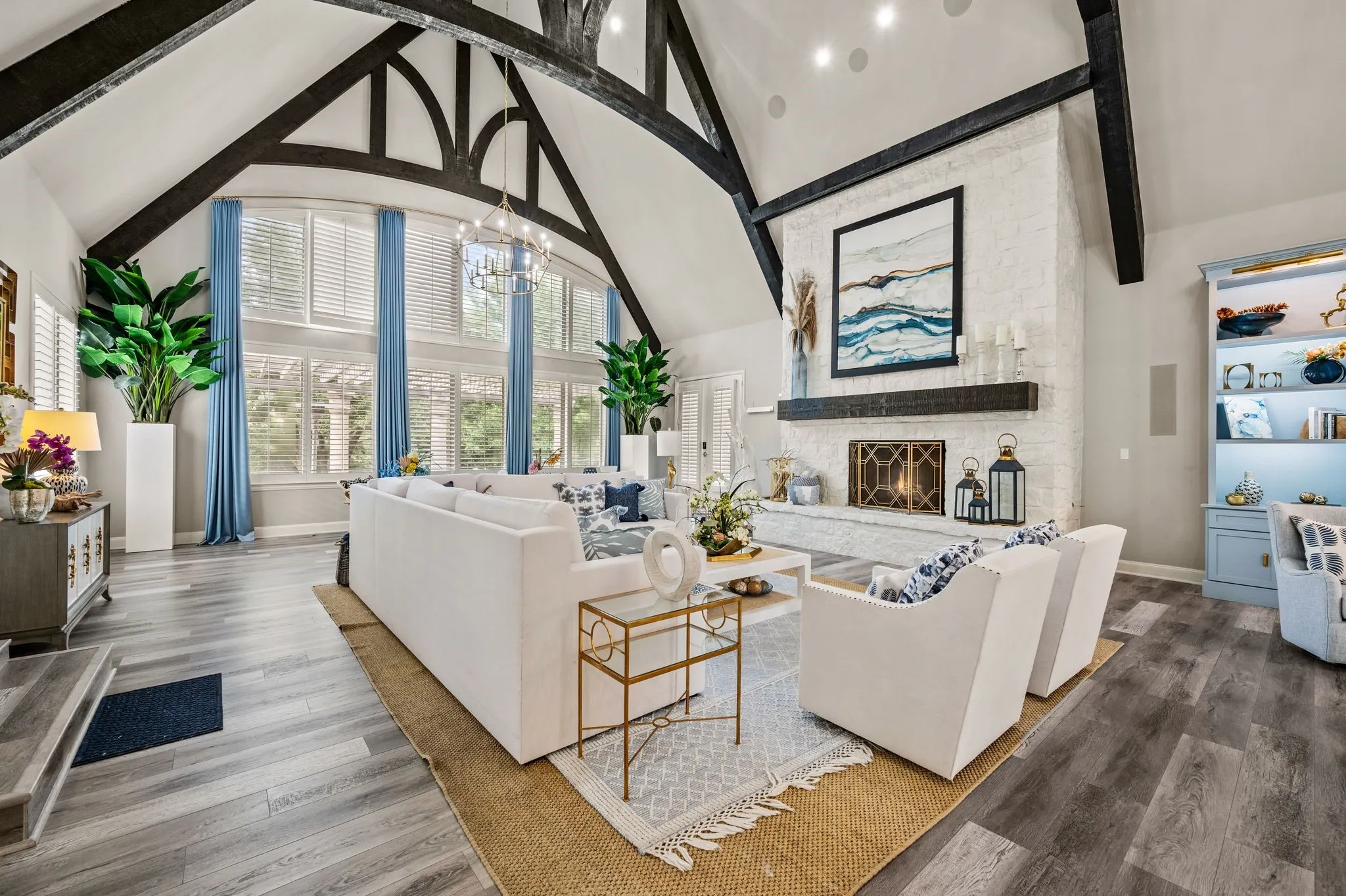 Vaulted ceiling with exposed wood beams, brick fireplace with wood mantel, wood-finish flooring, built-in shelving, and large windows with plantation shutters