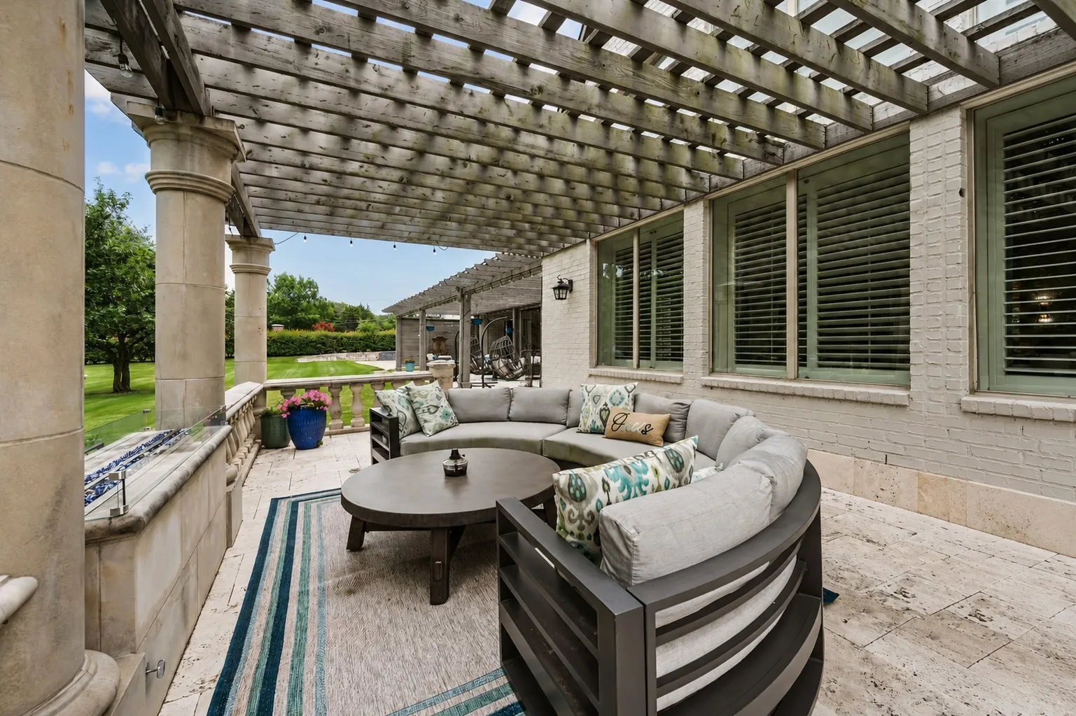 Expansive outdoor patio featuring a wood pergola, stone columns, a stone balustrade, brick exterior, and shuttered windows