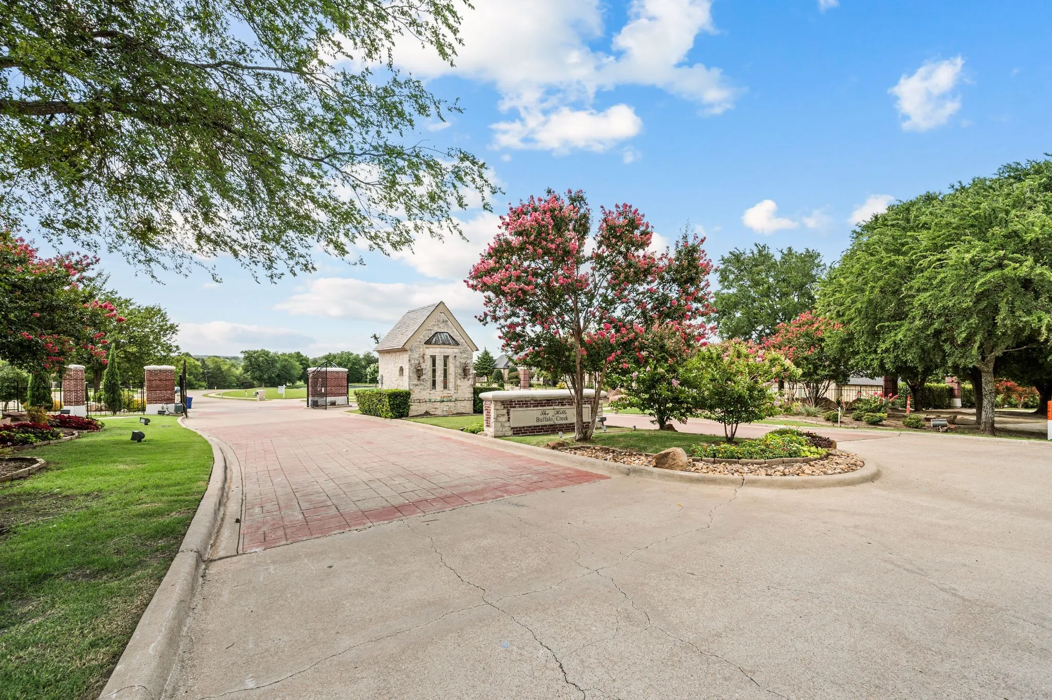 Entryway to the Hills of Buffalo Creek featuring a decorative brick-patterned drive, stone-clad gatehouse, and ornamental landscaping with mature trees