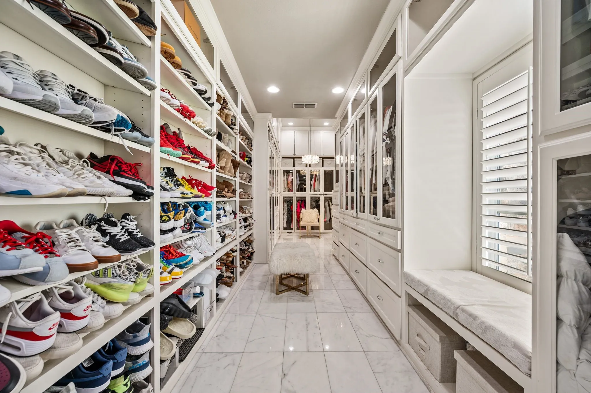 Custom closet featuring extensive open shelving, built-in cabinetry with glass-front doors, marble-look tile flooring, recessed lighting, and a window with plantation shutters